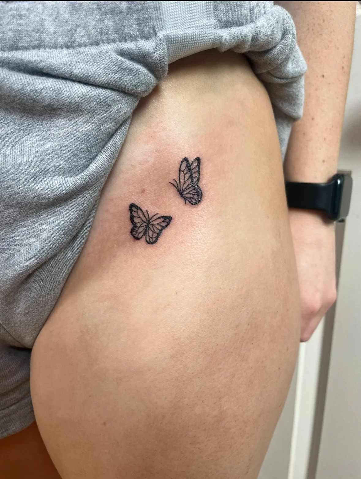 Close-up of a person's thigh with a black ink butterfly tattoo near the hip, partially covered by a gray shirt, with a black smartwatch on the wrist.
