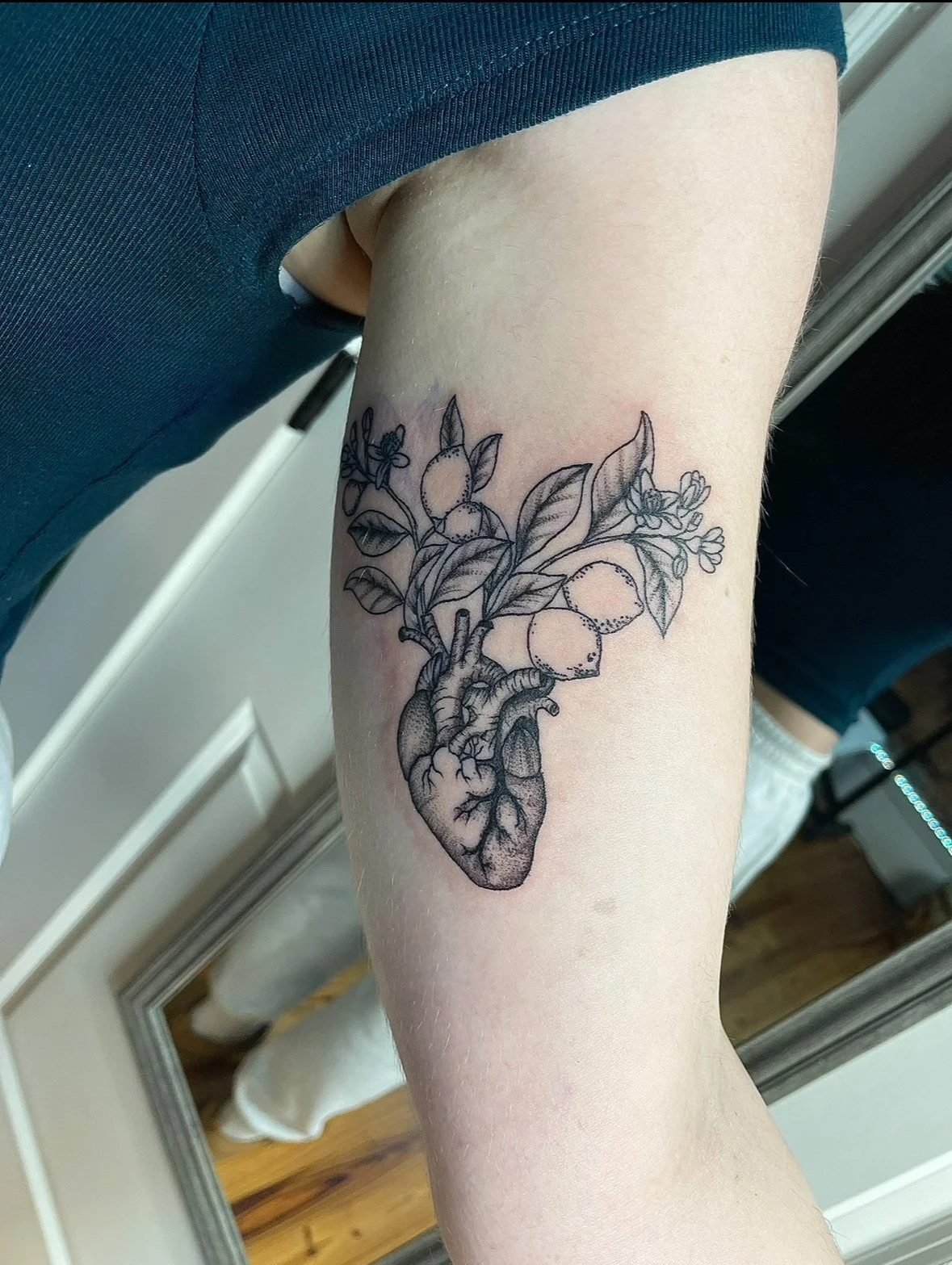 Black ink tattoo of a human heart with leaves, fruit, and small flowers growing from it, on a person's forearm.