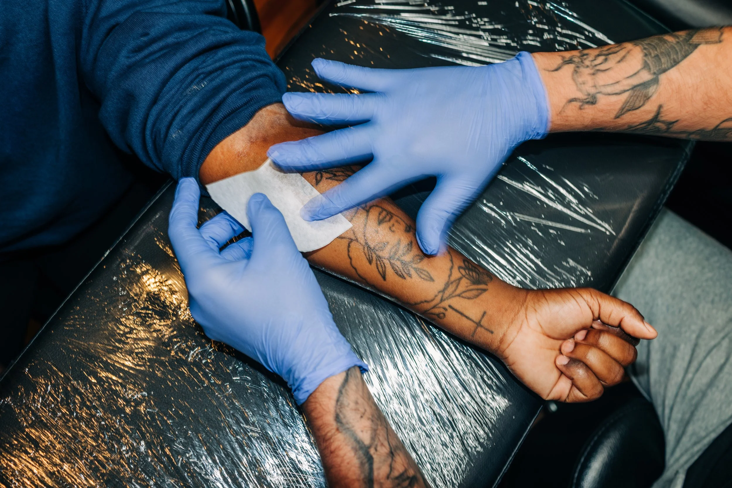 Tattoo artist wearing blue gloves applying ink to a person's arm, with tattoos visible on both arms, in a tattoo studio.