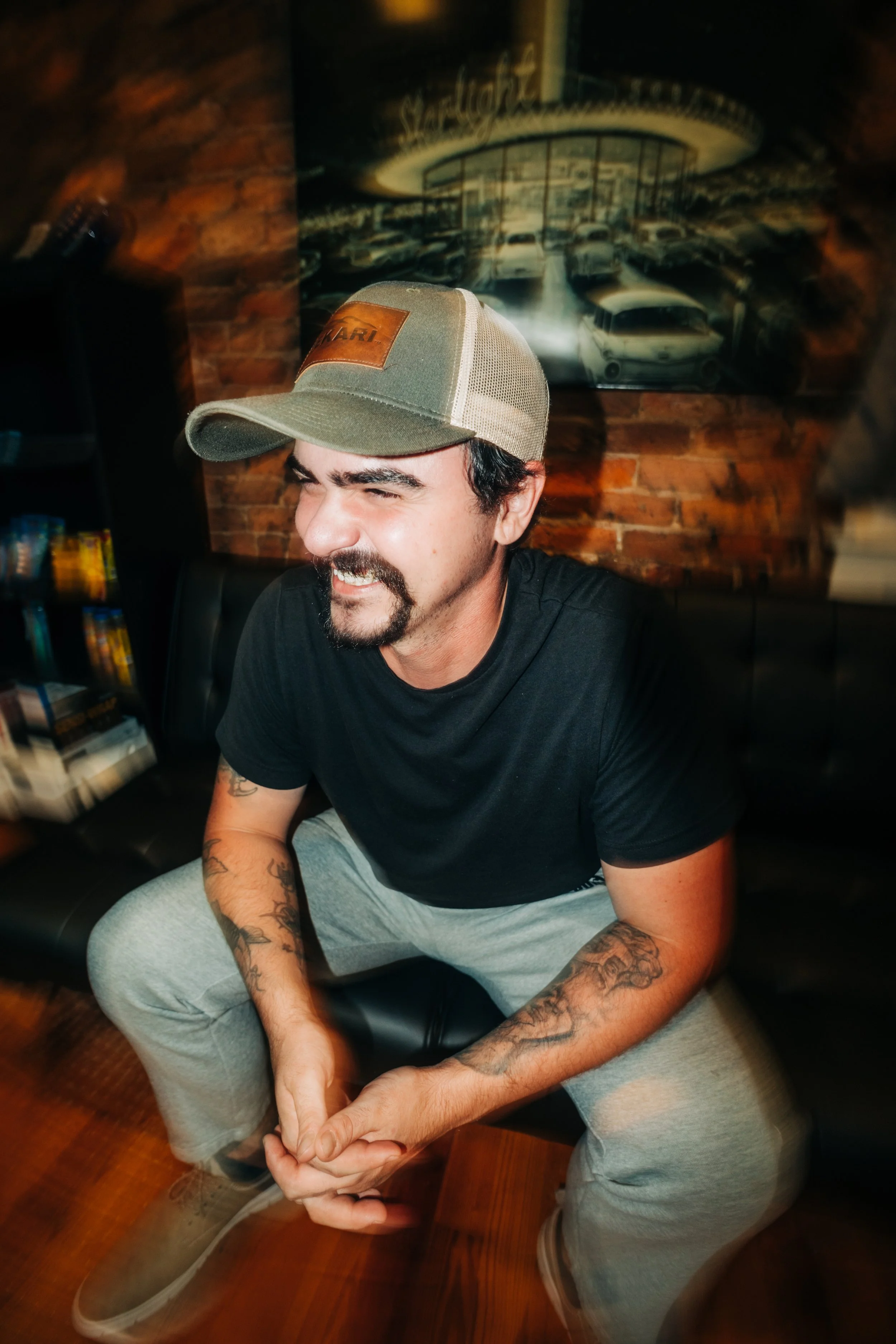 A man wearing a baseball cap and black t-shirt is sitting on a black leather couch in a room with an exposed brick wall. He is smiling with his eyes closed and hands clasped. There is a painting of classic cars and a neon sign behind him.