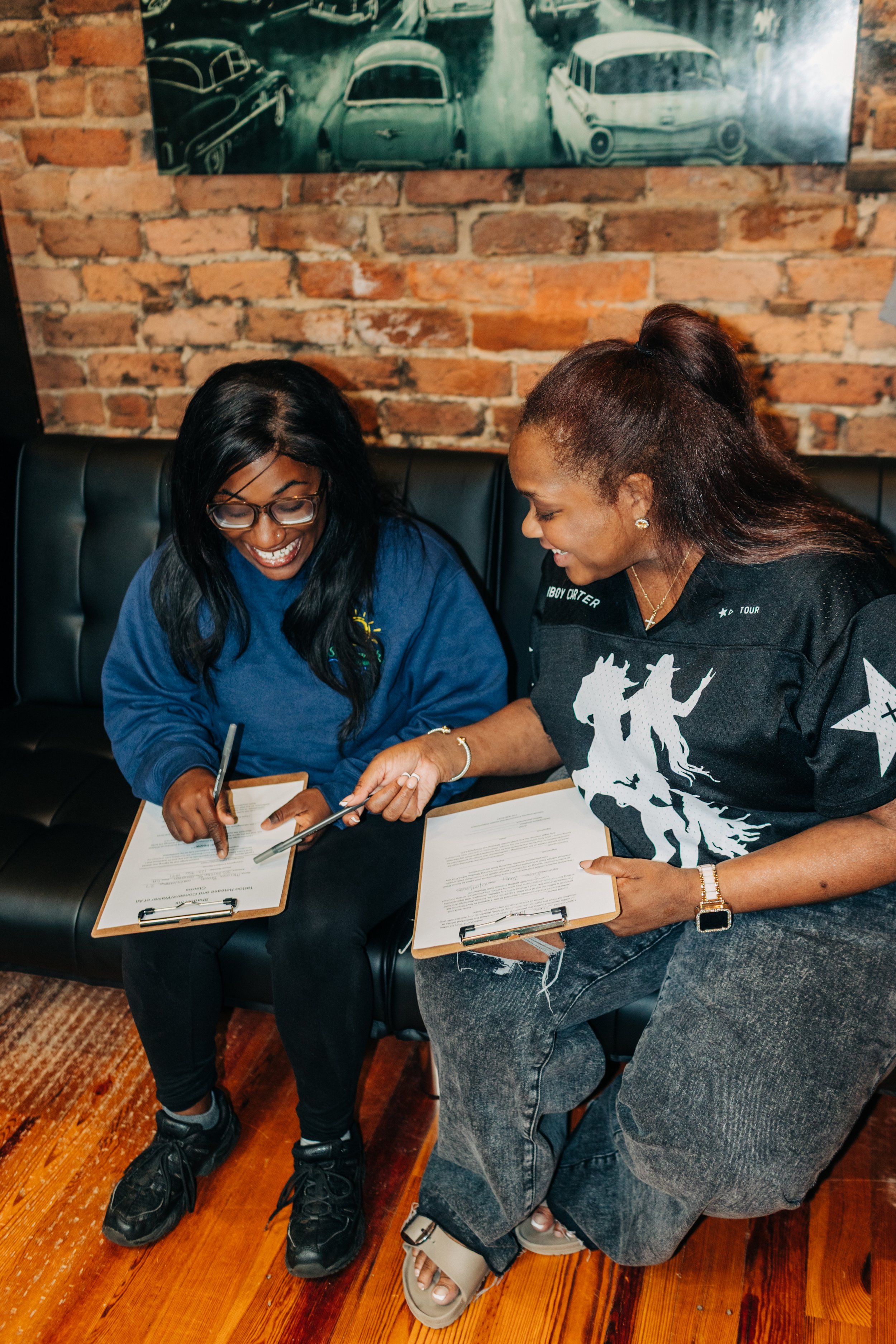 Two women sitting on a black leather couch, smiling and looking at documents on clipboards they are holding, in front of a brick wall with a vintage car-themed painting or photograph hanging on it.