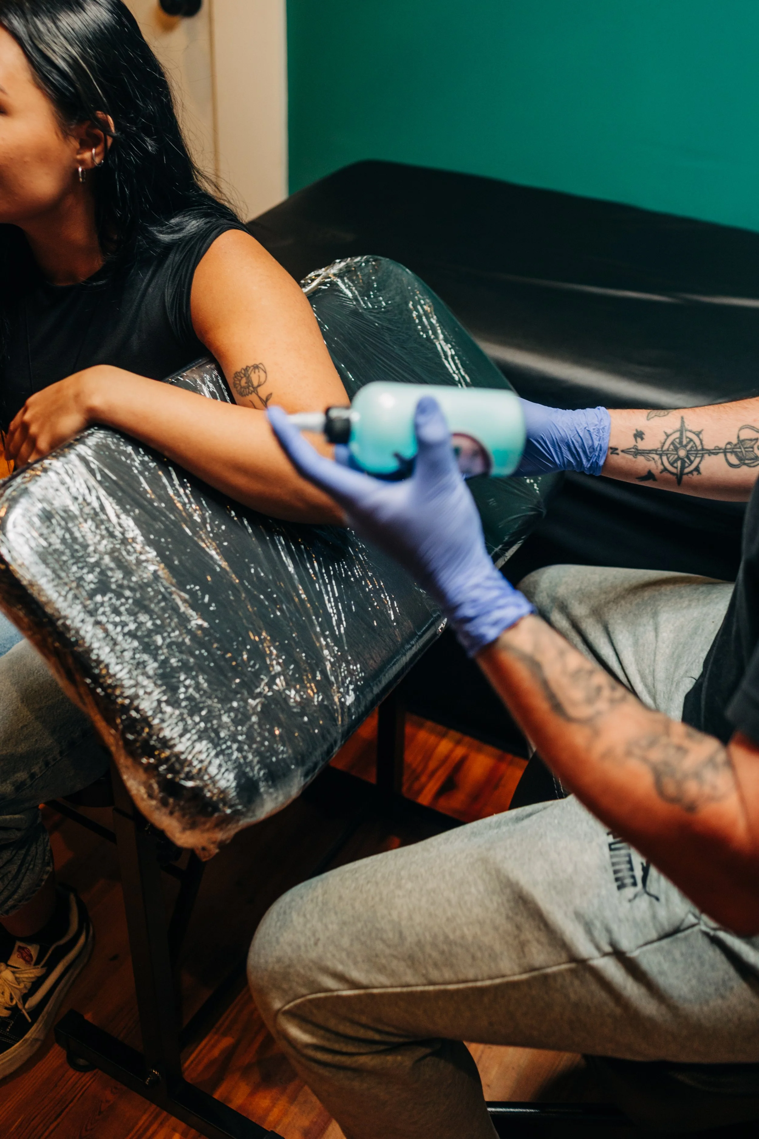A person getting a tattoo on their upper arm while seated in a tattoo chair. The tattoo artist is holding a tattoo machine, wearing blue gloves, and has tattoos on their arm. The person receiving the tattoo has dark hair, earrings, and a tattoo on their arm.