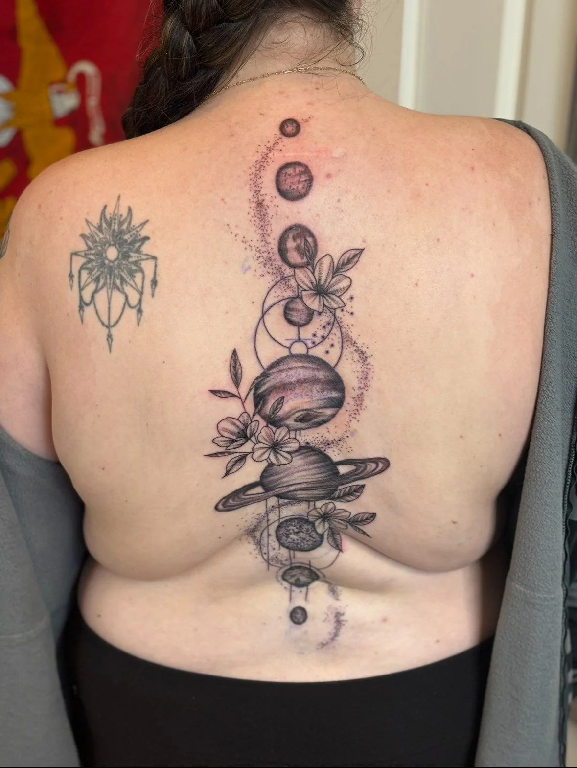 Tattoo of planets, flowers, and geometric designs on a person's back, with a smaller tattoo of a symmetrical, star-like design on the left shoulder.