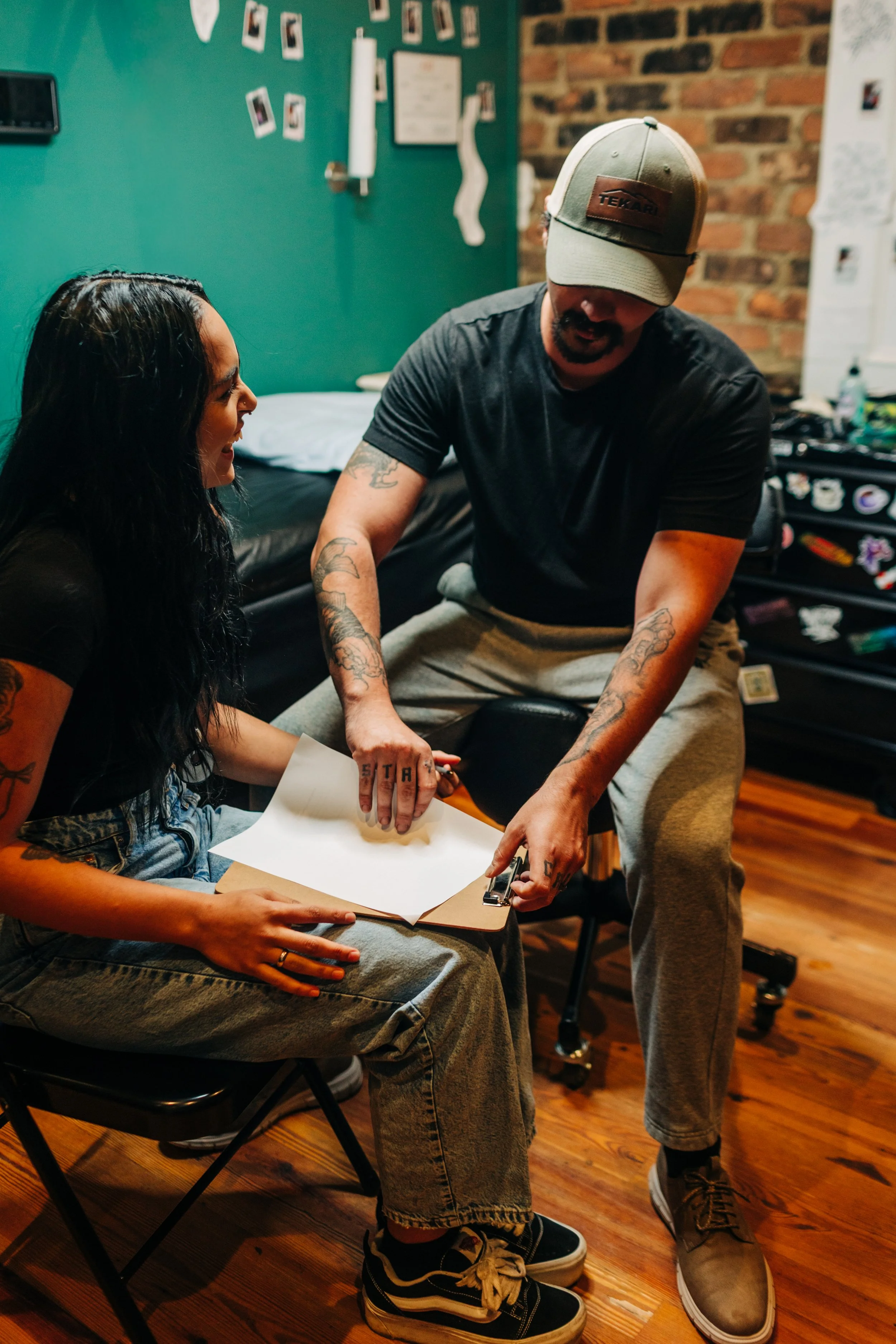 A woman sitting on a black chair showing an open book or notebook to a man. The man is standing, leaning over, and using a clip to attach papers in the notebook. They are in a room with a teal wall decorated with pictures and a brick wall. The woman has long black hair, tattoos, and is wearing ripped jeans and sneakers. The man is wearing a baseball cap, black t-shirt, gray sweatpants, and boots.