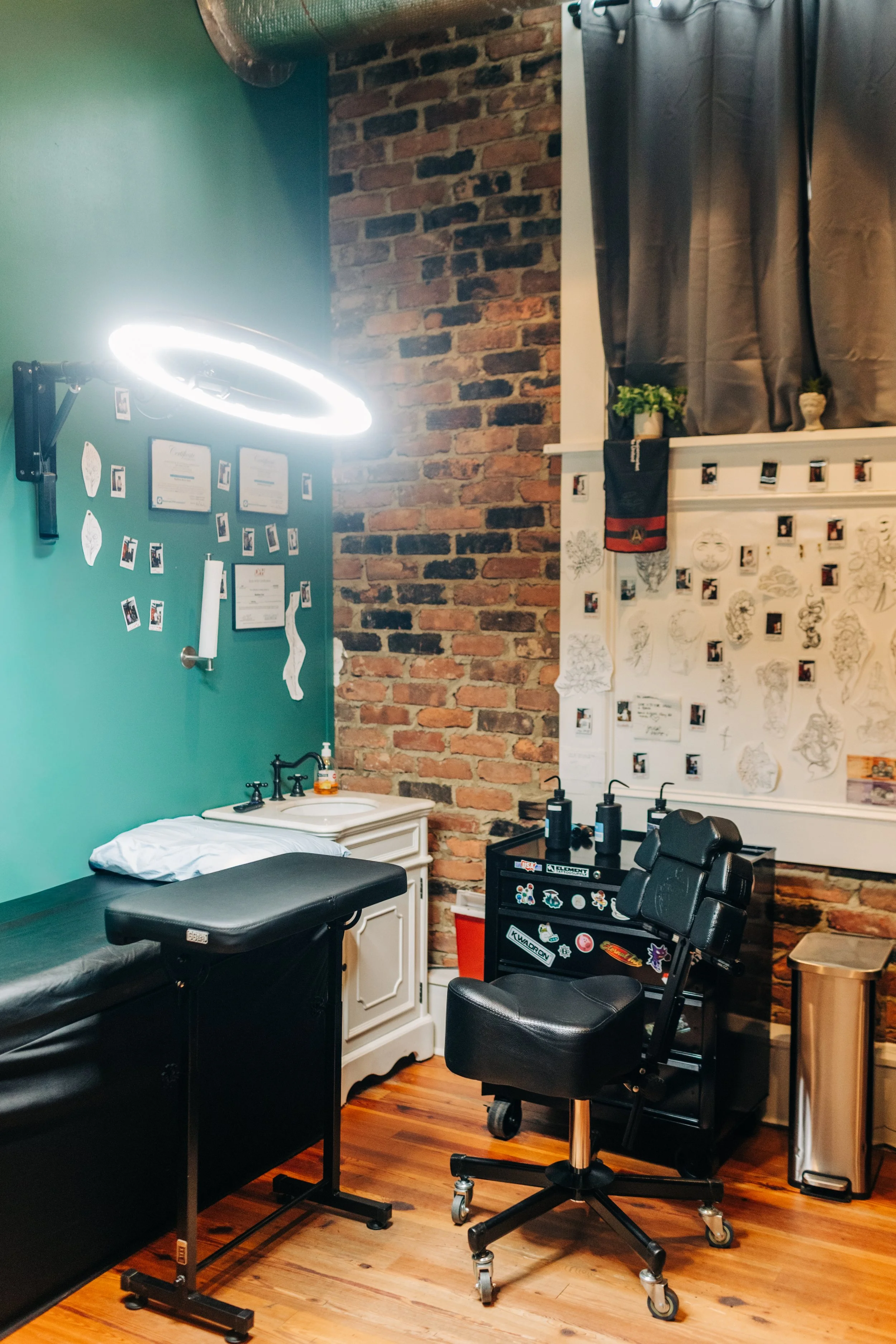 Tattoo studio room with a tattoo chair, a white sink, tattoo supplies, framed certificates on a teal wall, and a wall covered with tattoo sketches and Polaroid photos, in an interior with exposed brick wall and hardwood floor.