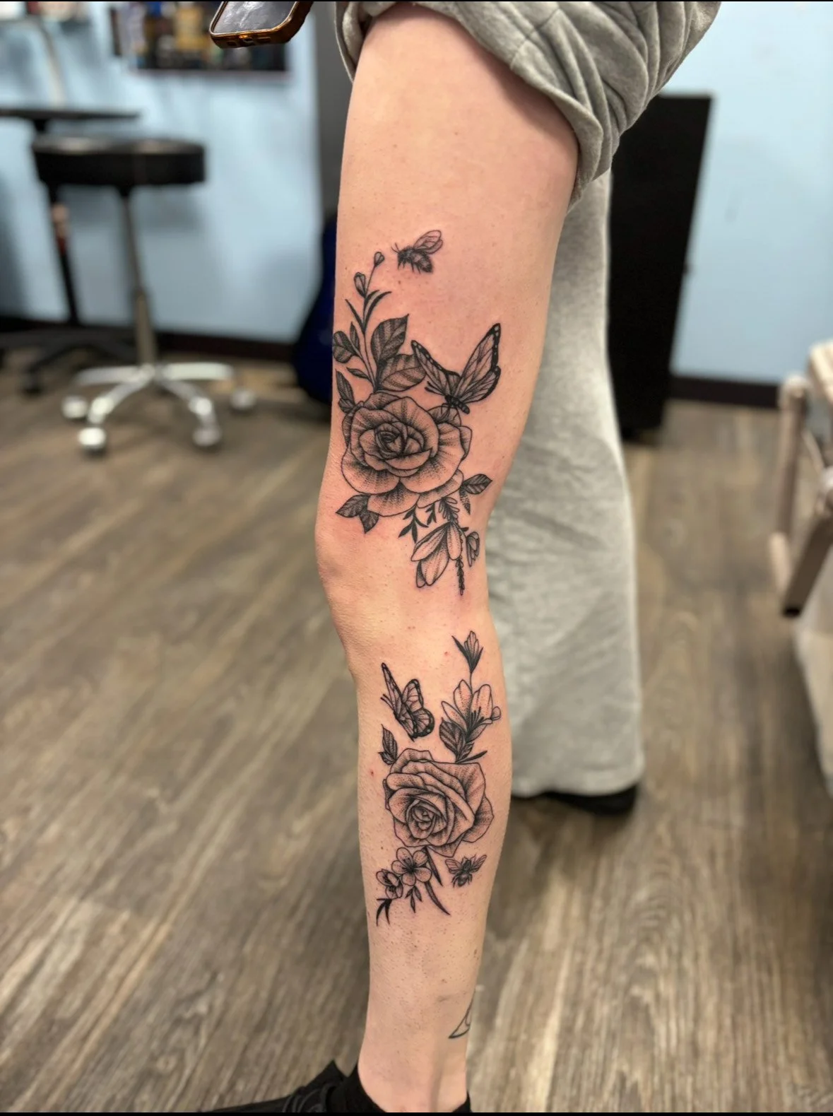 Black and gray floral tattoo featuring roses, butterflies, and leaves on a person's forearm.