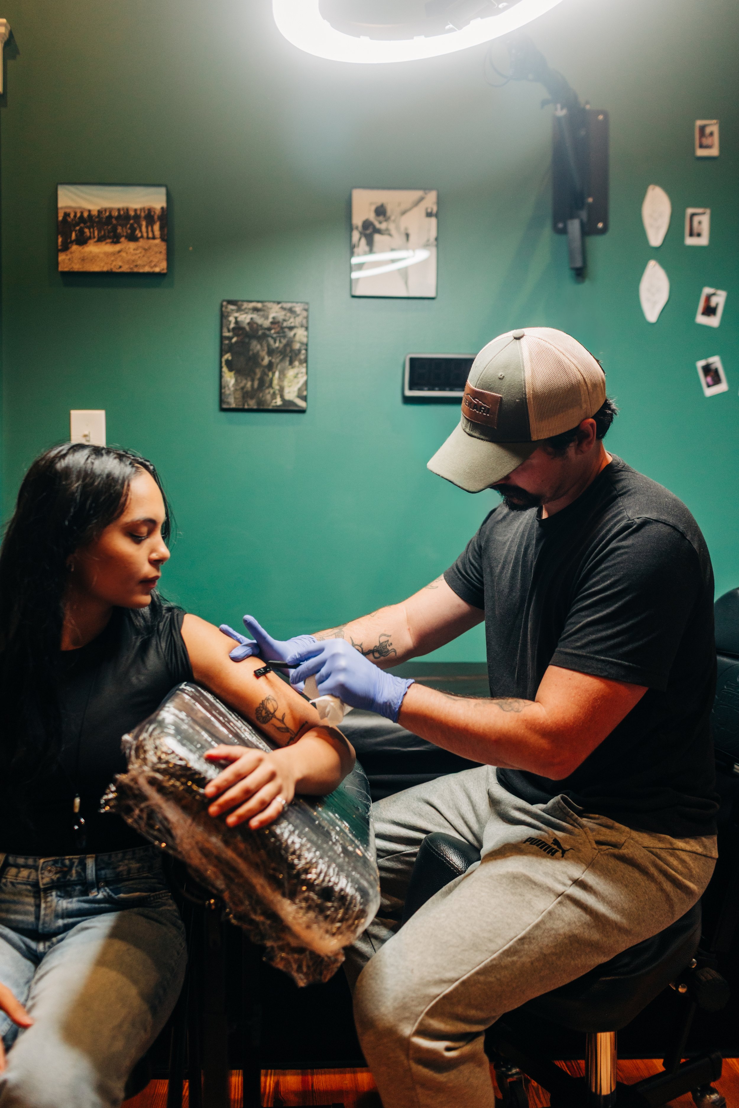 A woman receives a tattoo on her upper arm from a tattoo artist in a studio with dark green walls and framed photos and artwork. The woman is holding a sheet of shiny foil while the artist is wearing gloves and a cap.