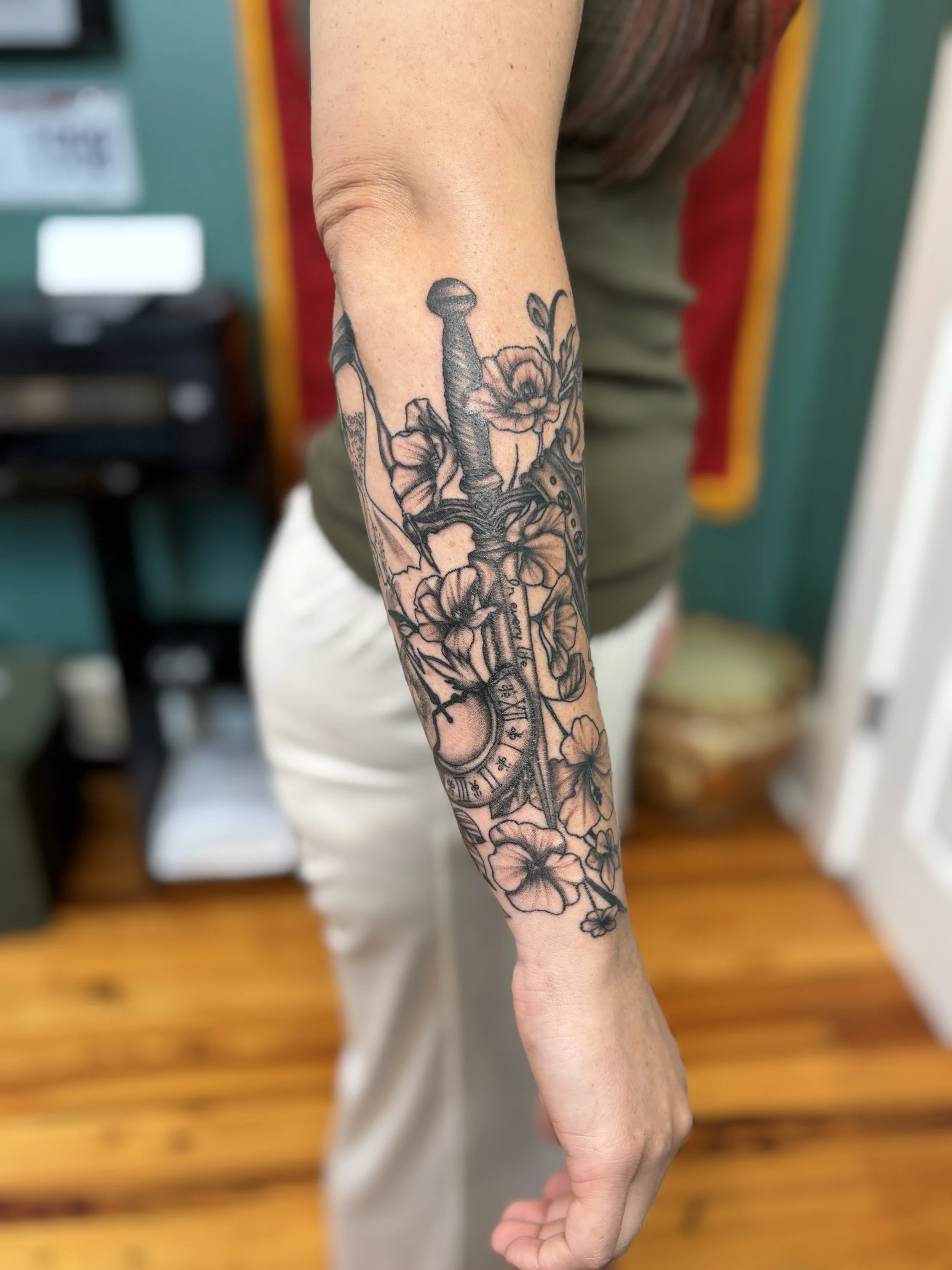 Black and gray tattoo on person's forearm featuring a sword, flowers, and a pocket watch.