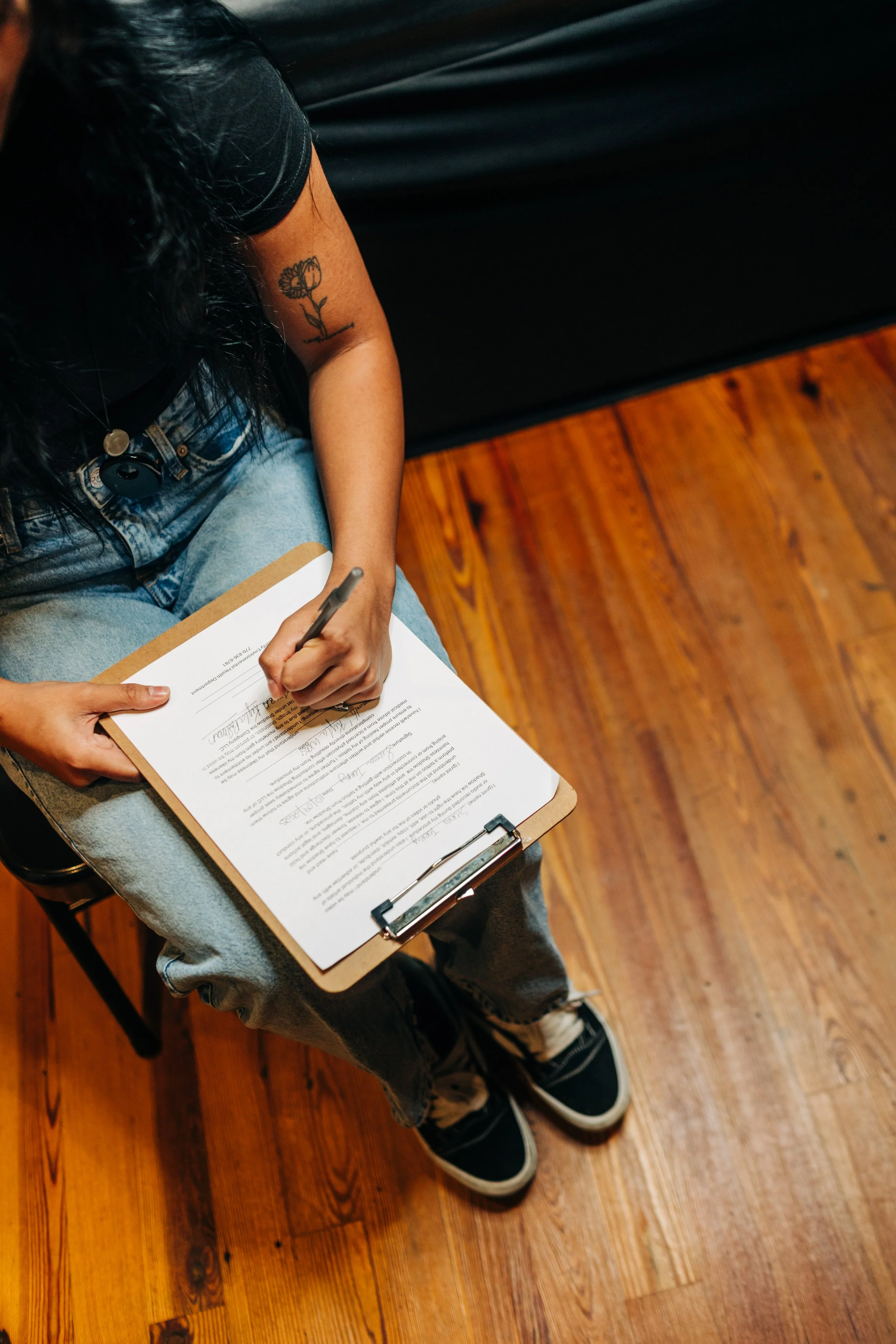 Person sitting on a chair, filling out a form or document with a pen on a clipboard, wearing casual clothing including jeans and sneakers, with a tattoo of a flower on their arm, on a wooden floor.