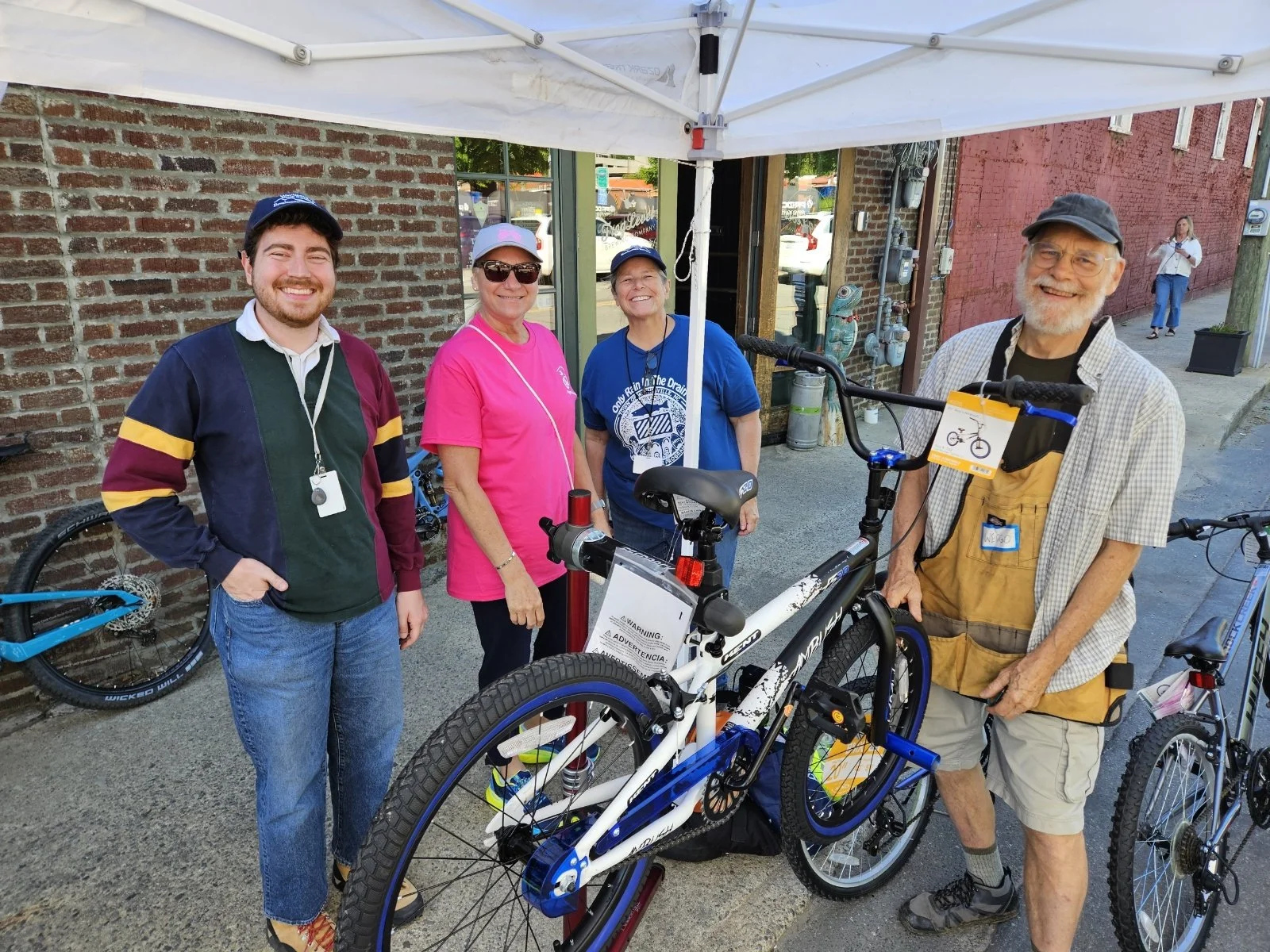 Four people standing under a white tent, smiling, with bicycles nearby, outside a brick building.