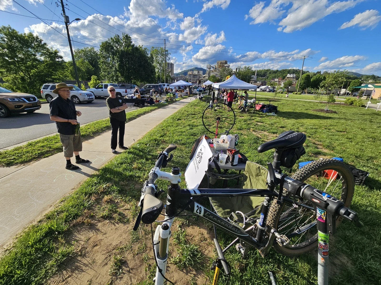 People at an outdoor market under blue sky with clouds, cars parked along the street, tents set up, and a bicycle parked in the foreground.