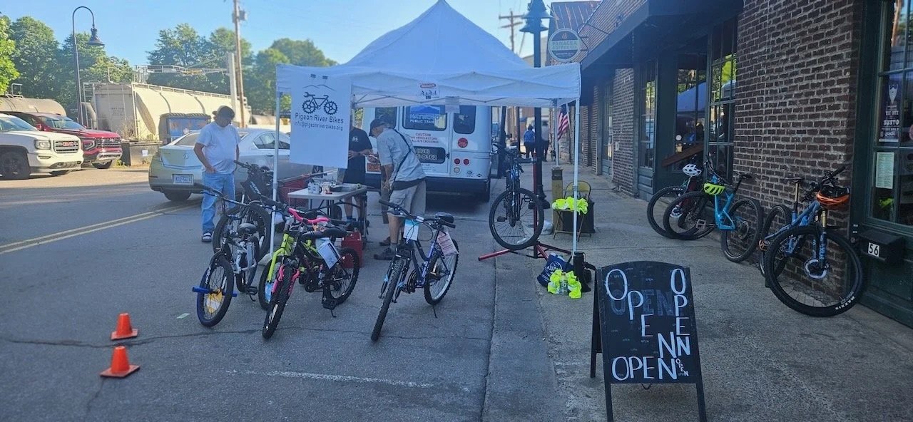 A bicycle shop stall outdoors on a sidewalk with several bikes, helmets, and accessories displayed. There are a few people talking, and a sign that says 'Open'. In the background, there are parked cars and buildings.