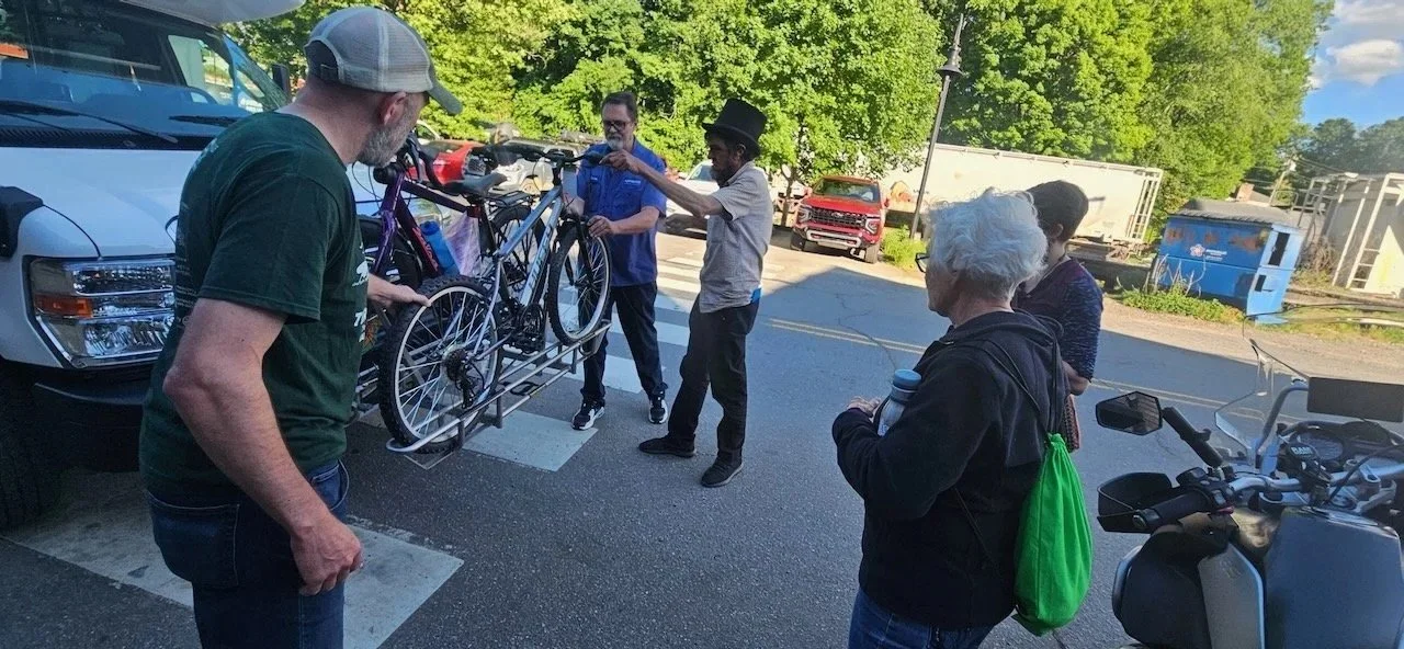 Group of five people gathered around a bicycle mounted on a vehicle, with trees and parked vehicles in the background.
