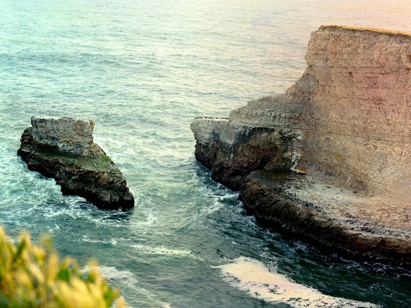 Cliffs along the ocean with waves crashing against the rocks, viewed during sunset.