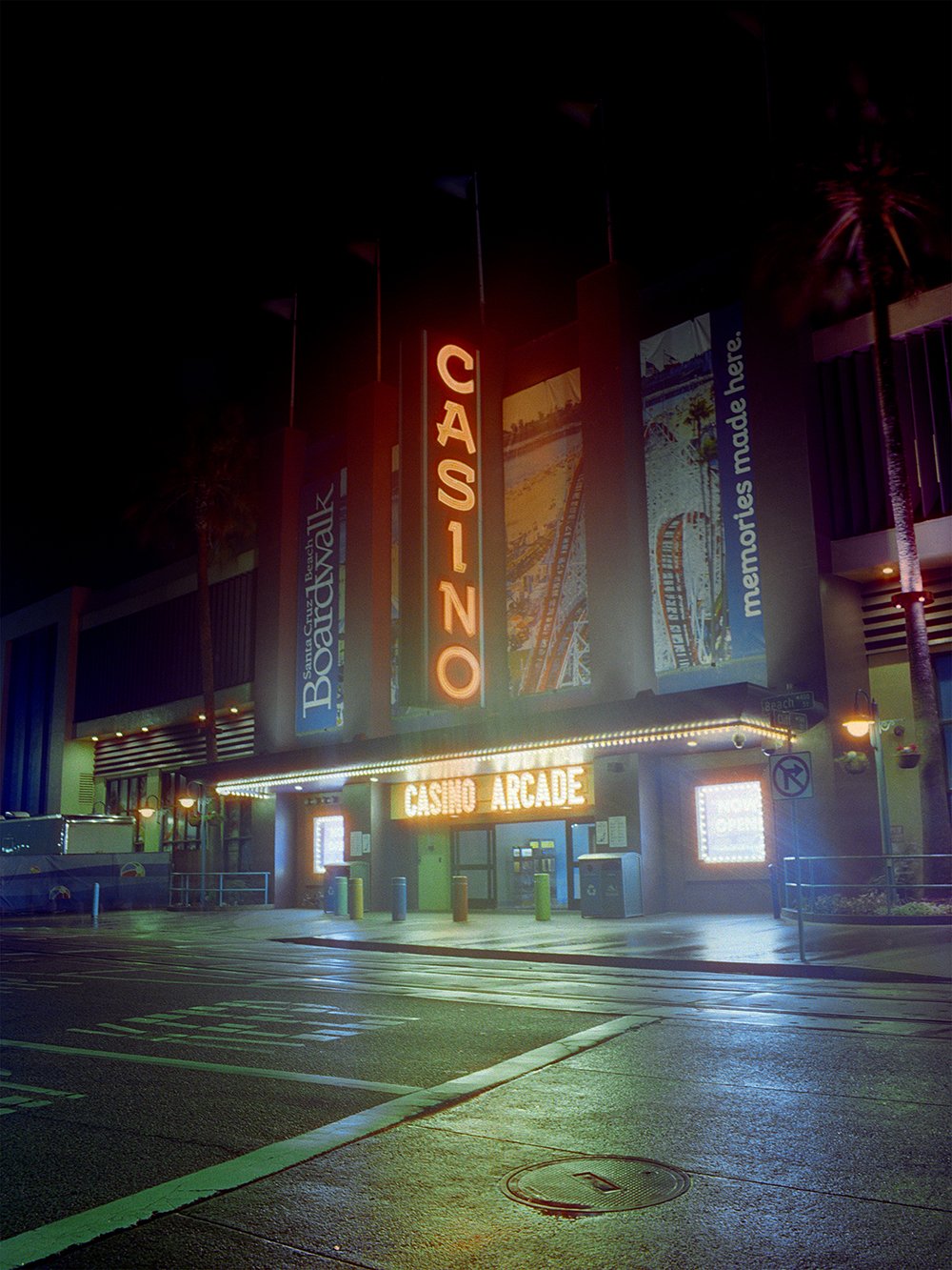 Night view of a casino arcade entrance with illuminated vertical sign displaying 'CASINO' and a marquee reading 'CASINO ARCADE' beneath it. The building is decorated with colorful banners and illuminated palm trees outside.