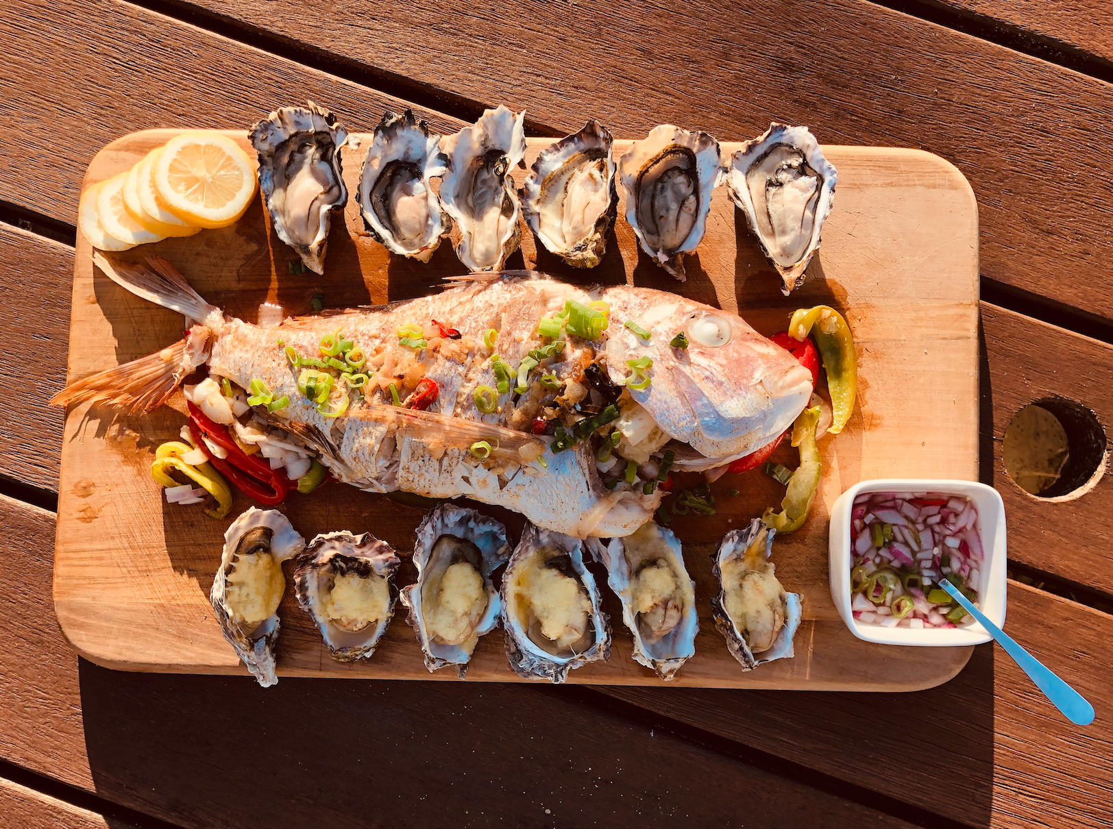 A wooden board with a whole grilled fish garnished with chopped green onions and sliced red and green peppers, around the fish are fresh oysters topped with butter, lemon slices, and a bowl of sliced red onions in vinegar. The setup is on a wooden table with sunlight.