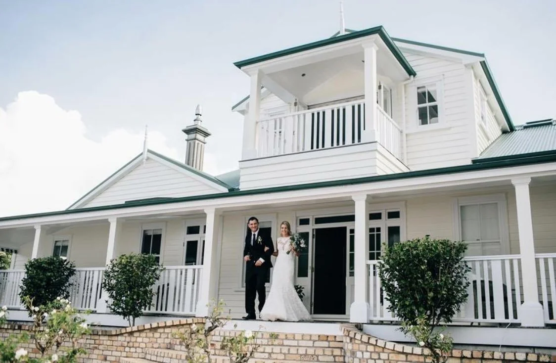 A bride and groom standing on the porch of a white house with a green roof, surrounded by small bushes and plants.