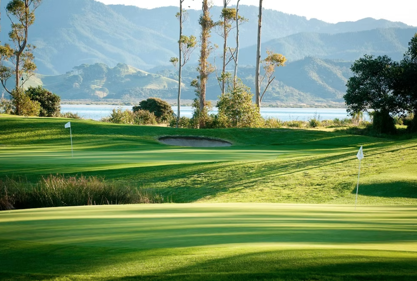 A beautiful golf course with green grass, flags on the putting greens, sand trap, surrounded by trees with mountains and water in the background.