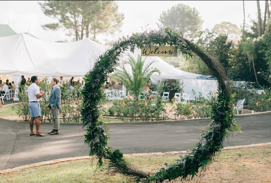 A wedding reception outdoors with a floral welcome arch, tents, and guests mingling.