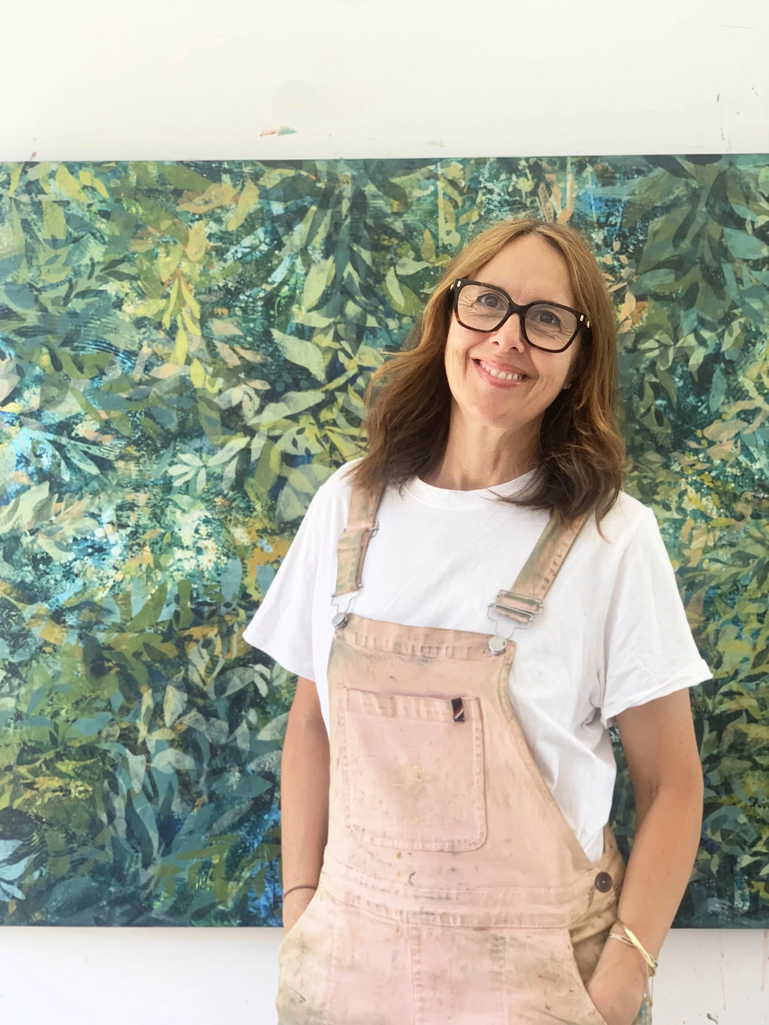 A woman with red hair, glasses, and a white t-shirt standing in front of a colorful abstract leafy painting, smiling.