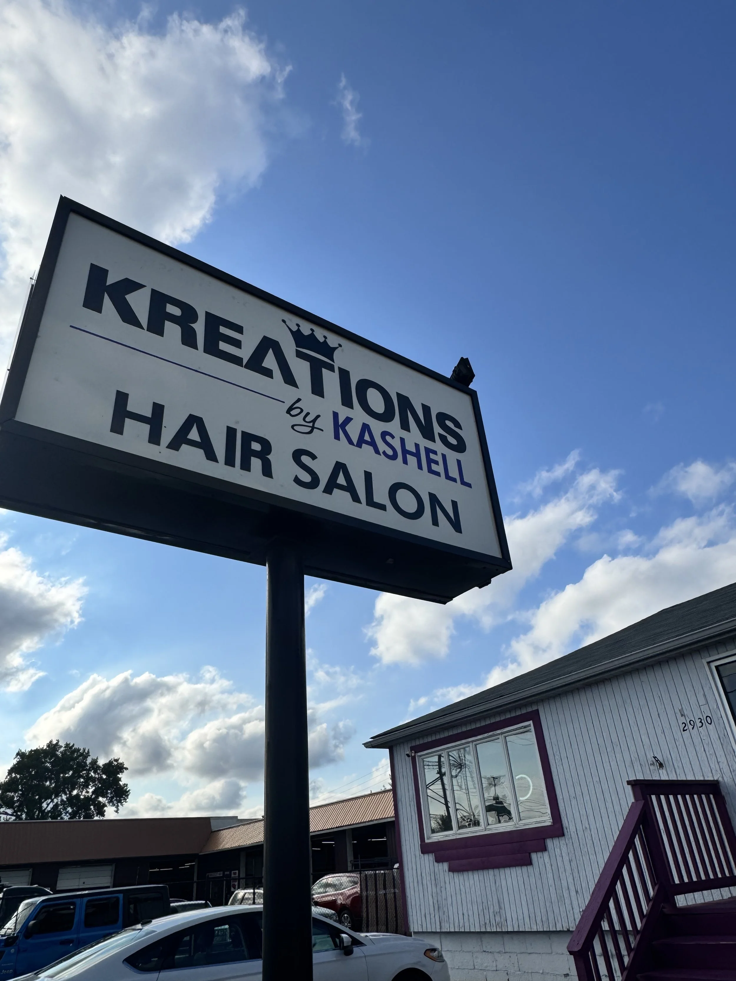 Sign for Kreations by Kashell Hair Salon against a partly cloudy sky with buildings and parked cars visible.