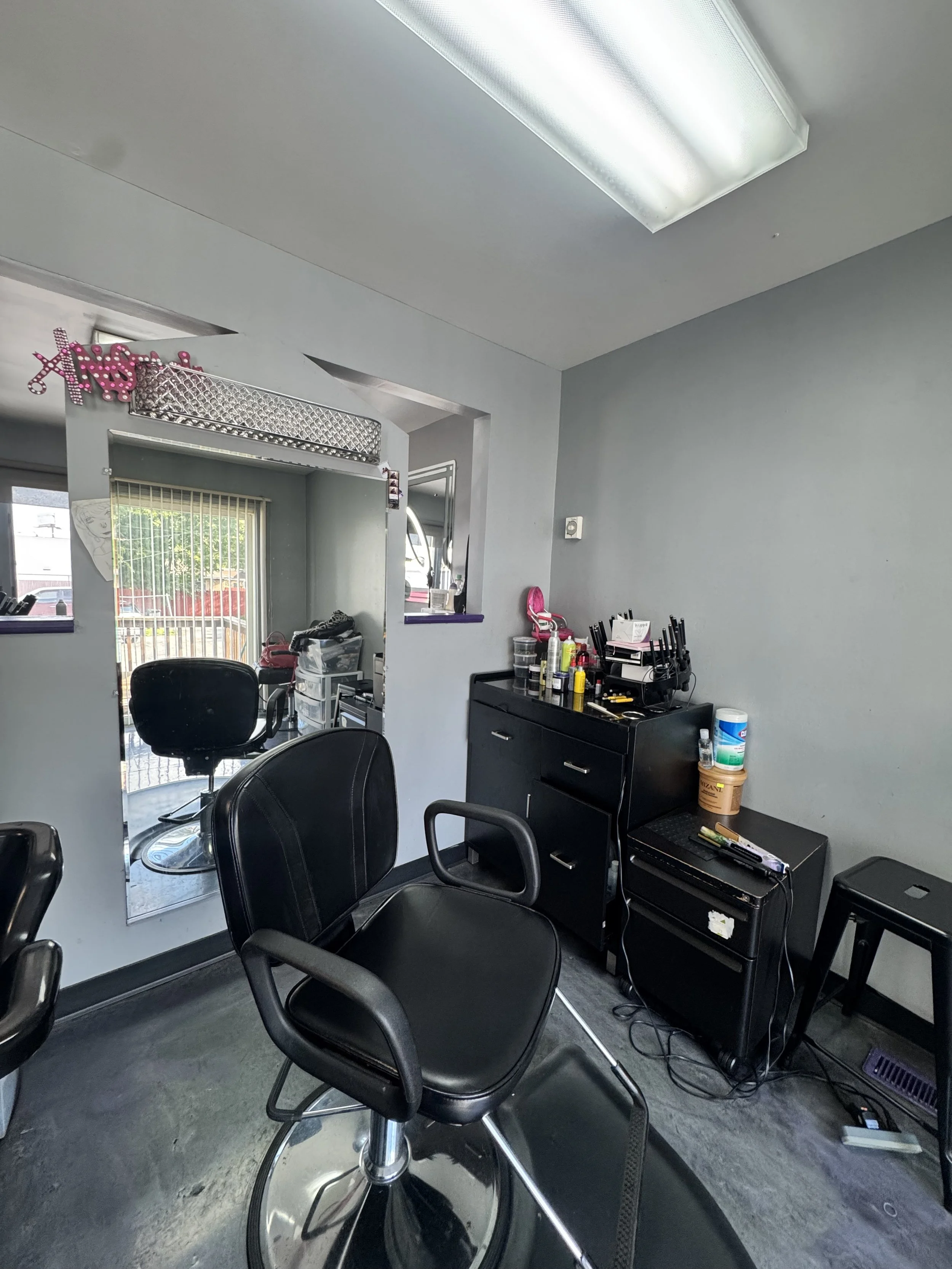 Interior of a hair salon or barbershop with black salon chairs, a black rolling cart with hair products and tools, and a mirror on the wall reflecting part of the room.