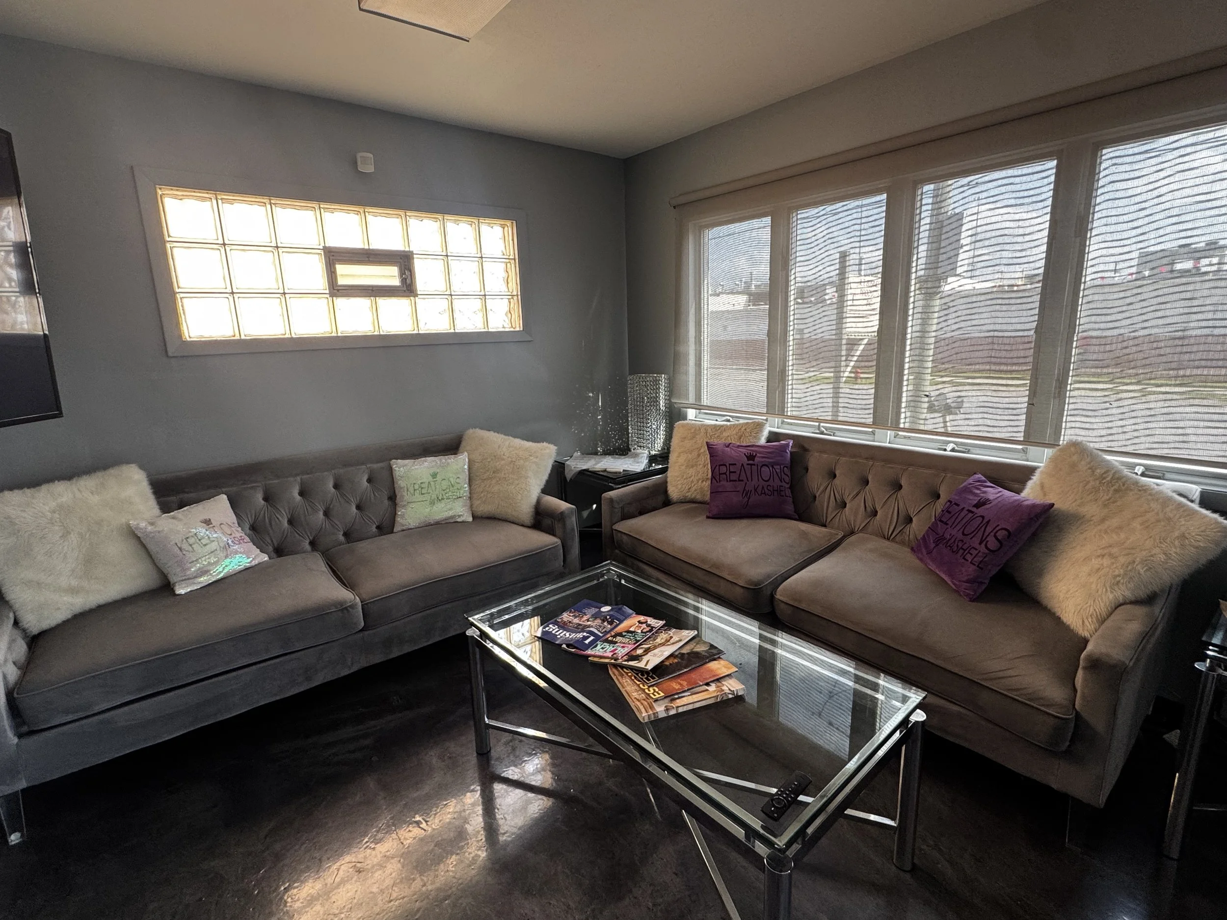 The Kreations by Kashell waiting area, couches decorated with cushions including fluffy pillow and branded pillows, and a glass coffee table, with large windows covered by blinds letting in natural light.