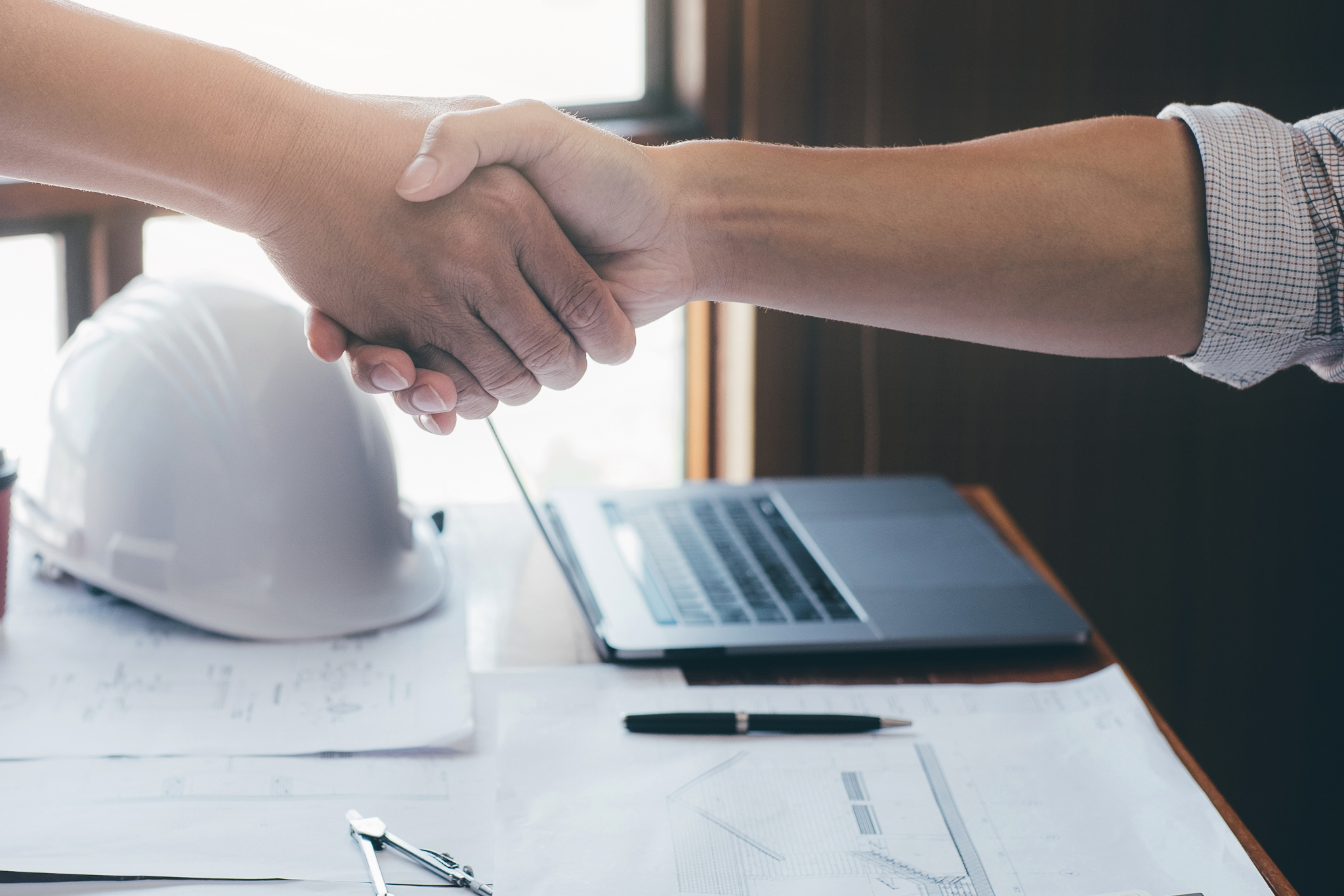 Construction professionals shake hands over project plans, representing the weekly financial reviews contractors use to monitor job costs, cash flow, change orders, and profitability.
