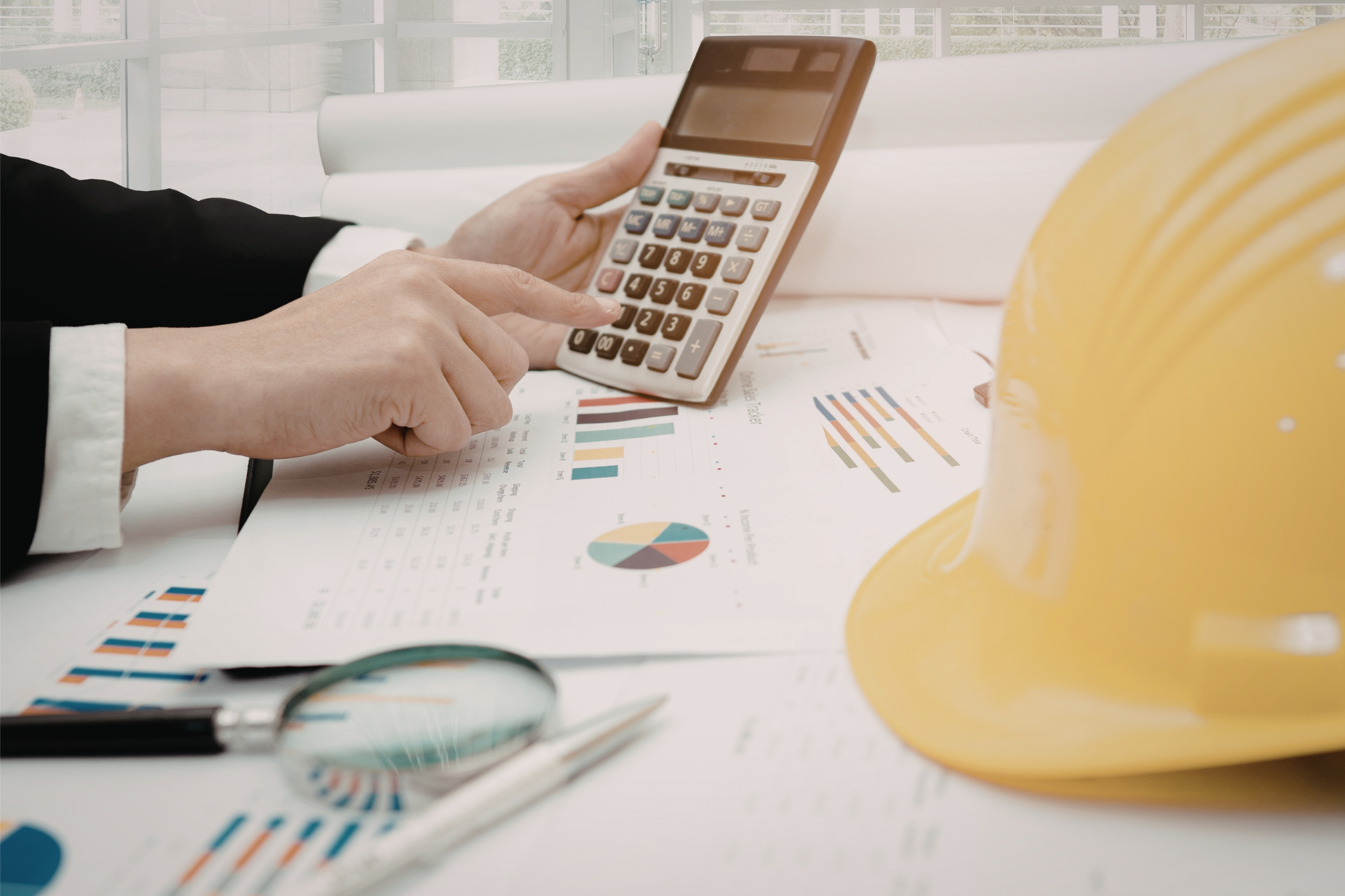 Hard hat, construction paperwork, and calculator on a work surface representing contractor cost tracking, highlighting the difference between fixed and variable costs and how they impact job profitability and overall business performance.