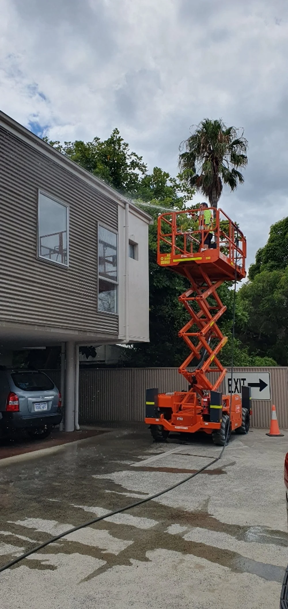 A person on an orange scissor lift washing or inspecting the exterior of a building with corrugated metal siding, with a palm tree and cloudy sky in the background.