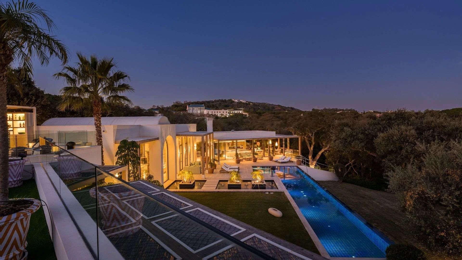 A modern house with outdoor pool area, lit up at dusk, surrounded by trees and hills in the background.