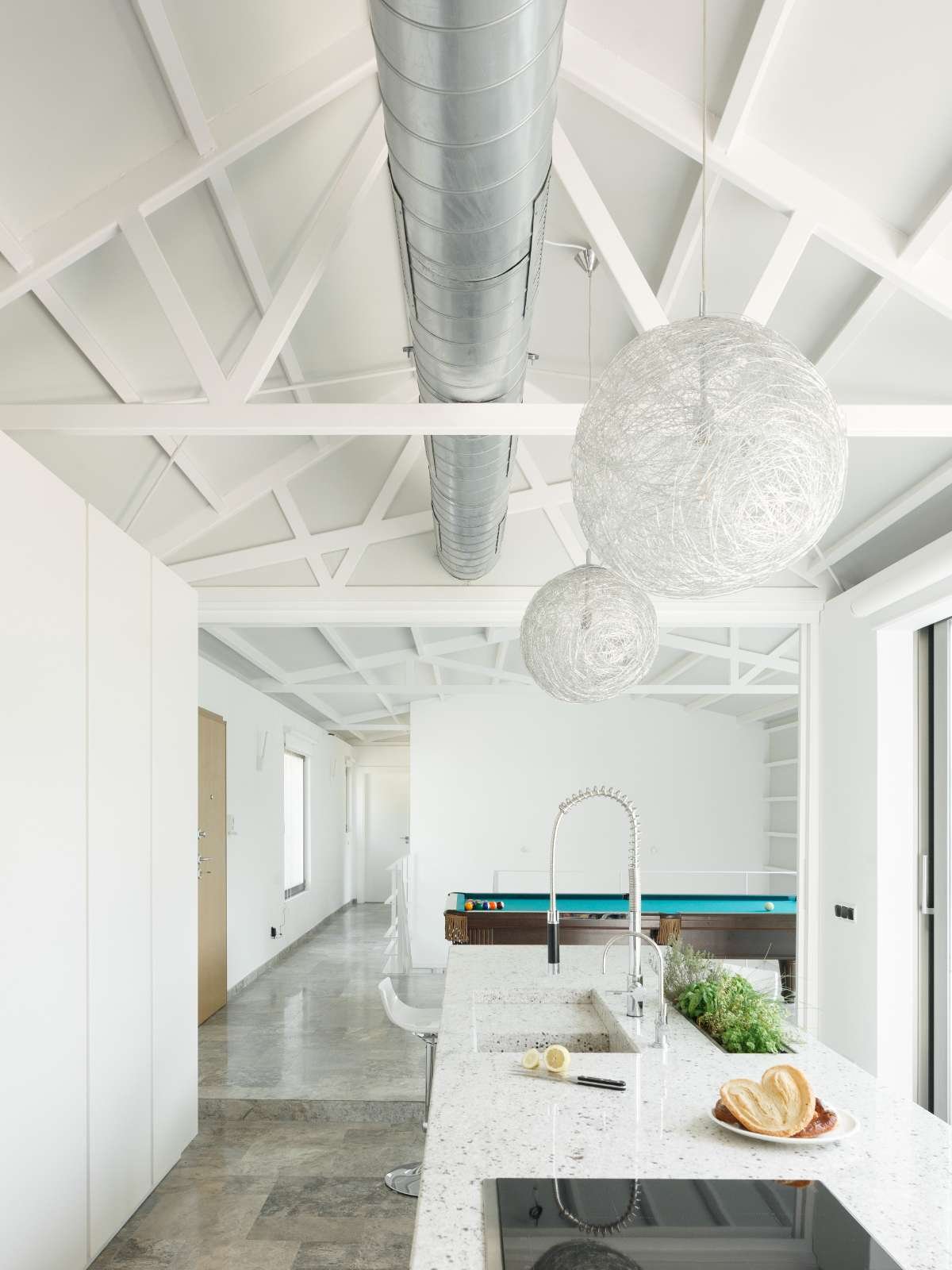 Modern kitchen with white cabinetry, a granite countertop, a sink with a flexible faucet, and hanging spherical pendant lights. A pool table is visible in the background, along with a hall with doors and windows.