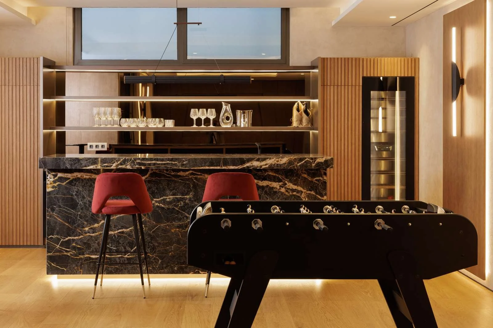 Modern home bar area with black marble countertop, red bar stools, a foosball table in the foreground, open shelves with glasses and decorative items, large window with a view outside, warm wood paneling, and ambient lighting.