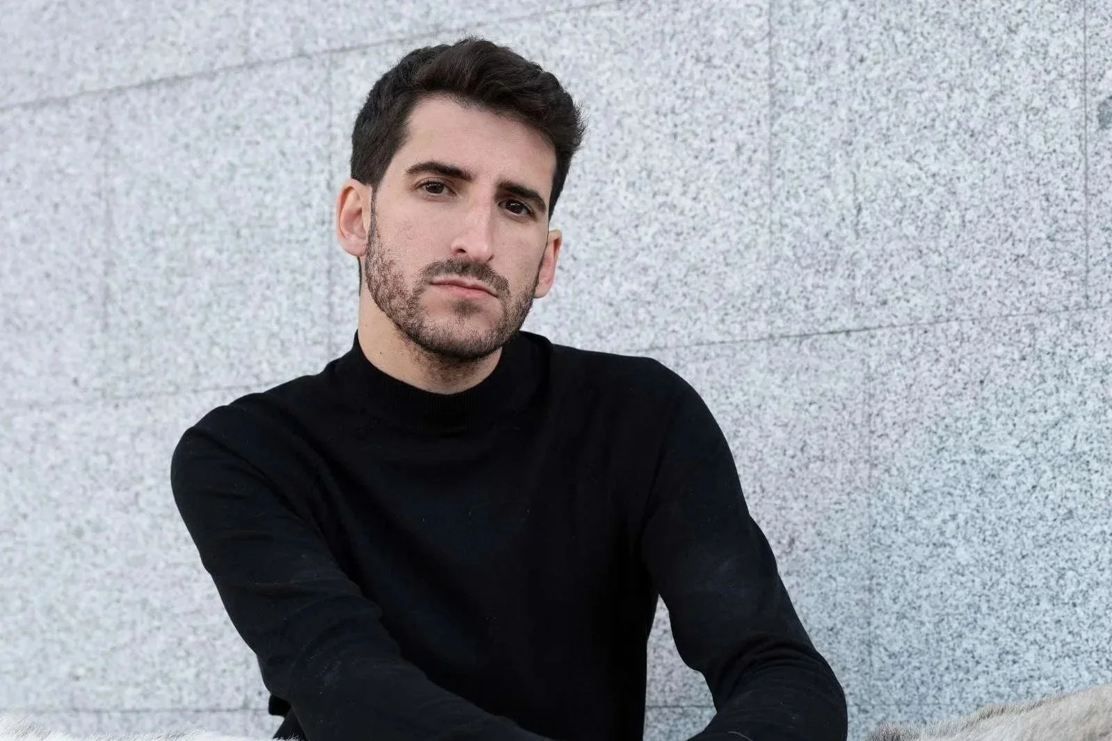 A man with dark hair and a beard wearing a black turtleneck sweater, sitting against a light gray textured wall, looking directly at the camera with a serious expression.