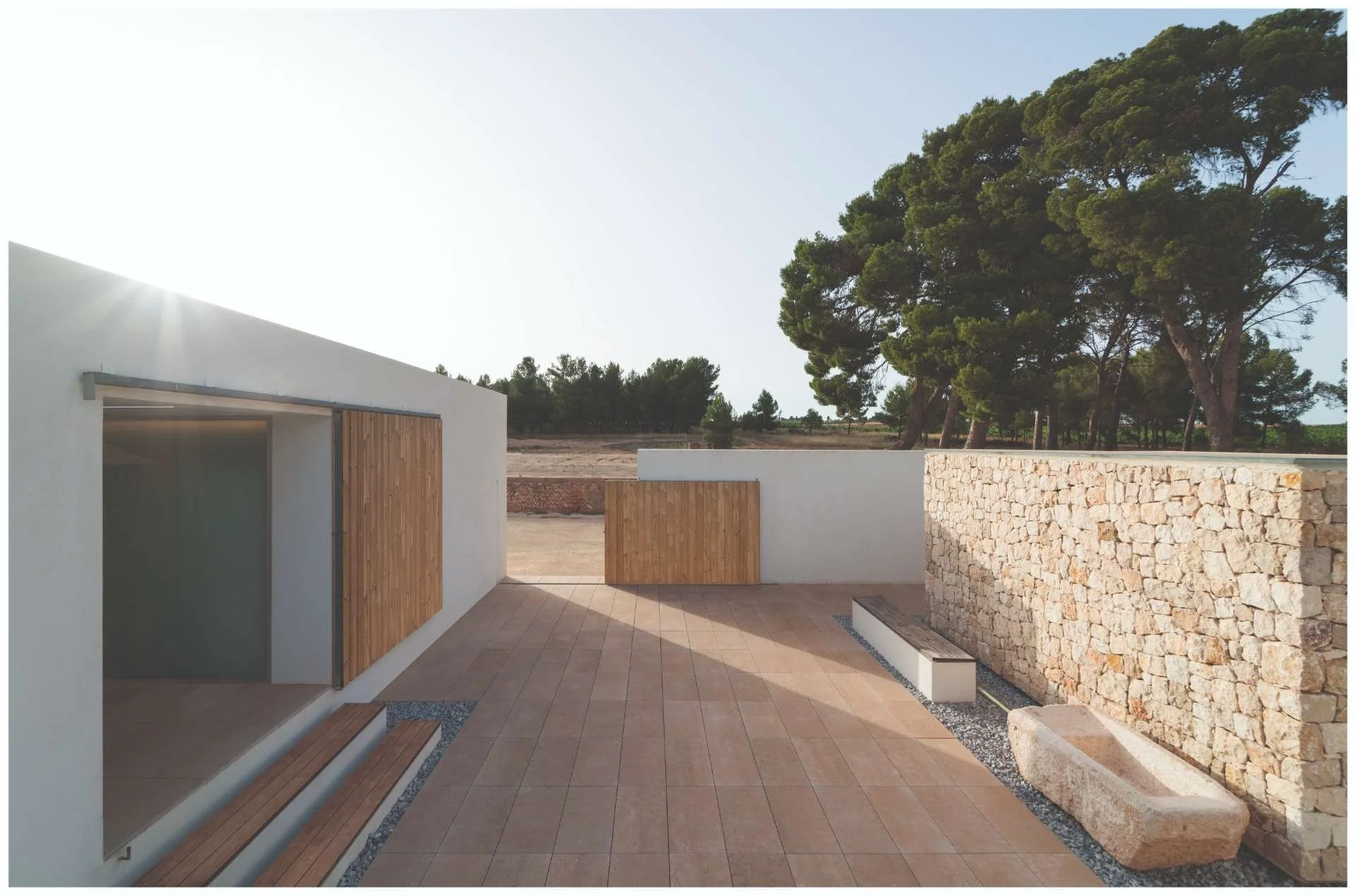 Modern outdoor patio area with a stone wall, tiled flooring, a wooden door, and surrounded by trees and open land