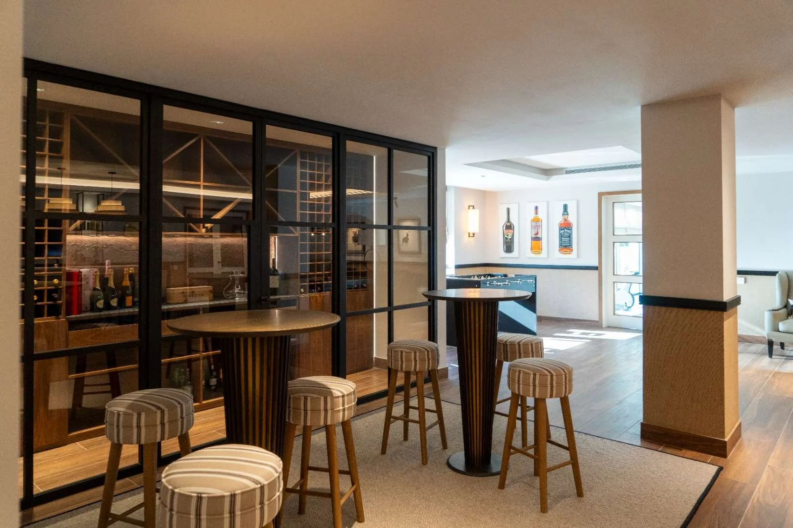 Interior of a bar with high table and stools, wine bottles behind glass partition, and framed liquor bottles on wall, with wooden floors and modern decor.