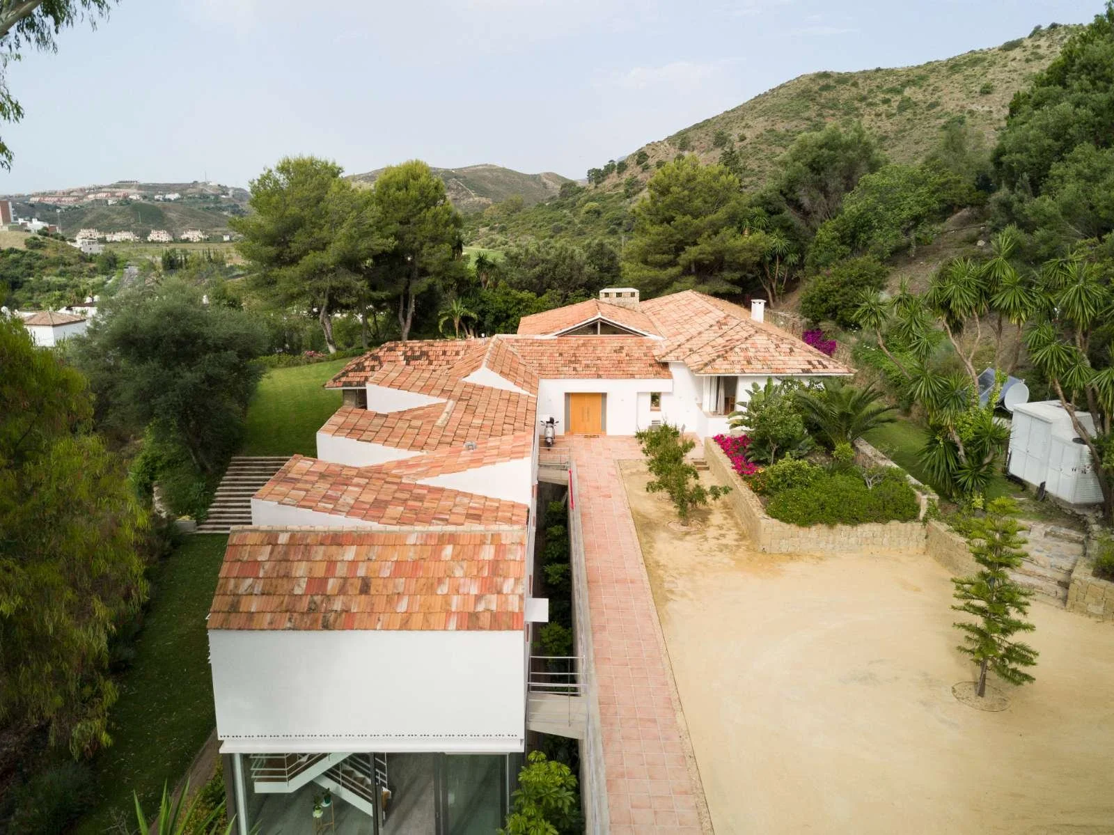 A house with white walls and red tile roof, surrounded by trees and greenery, with rolling hills in the background.