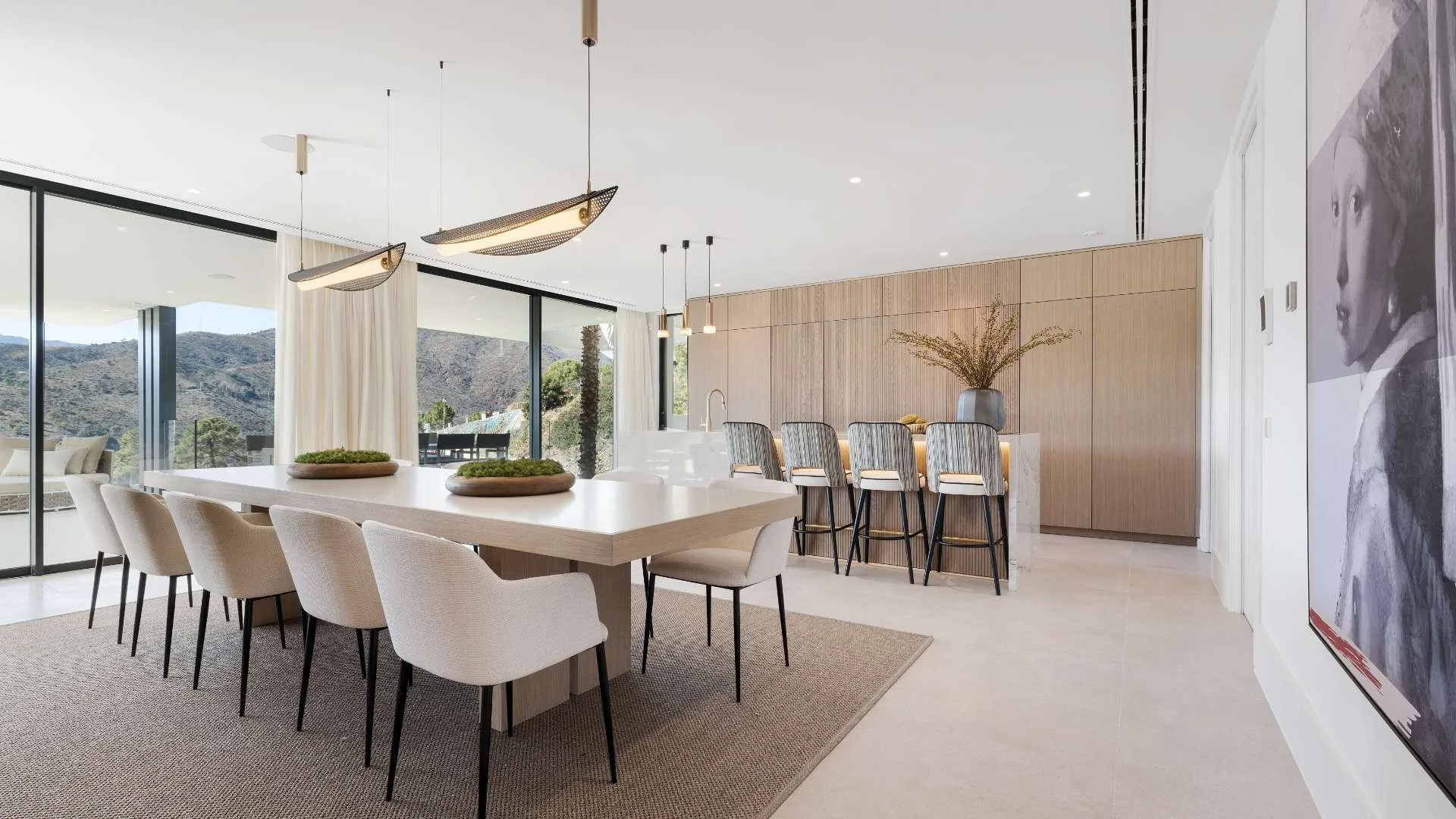 Modern dining area with a large rectangular table surrounded by light-colored upholstered chairs, pendant and ceiling lights, a bar-style counter with barstools, large windows with sheer curtains revealing outdoor mountains, a large vase with dried branches, and a black and white portrait on the wall.