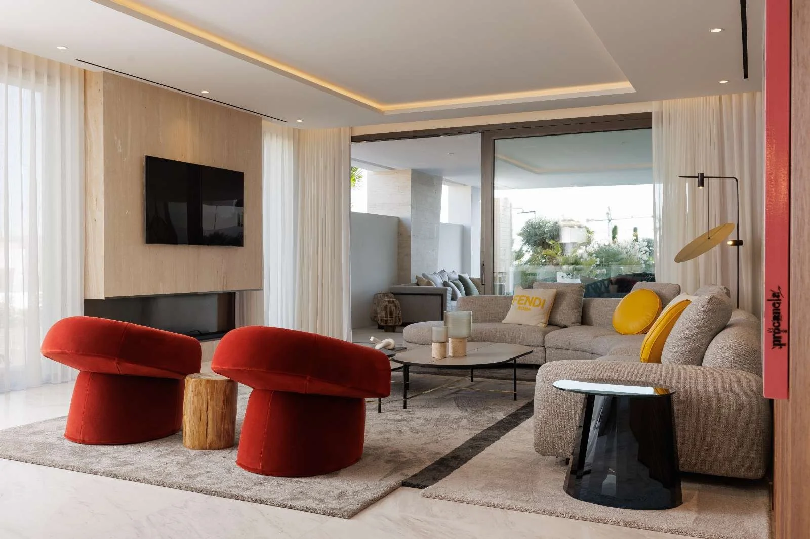 Modern living room with beige sofa, yellow accent pillows, and two red velvet chairs. A flat-screen TV is mounted on a beige wall, with large windows and sheer curtains letting in natural light. Coffee tables hold decorative items, and there is an outdoor patio visible through sliding glass doors.