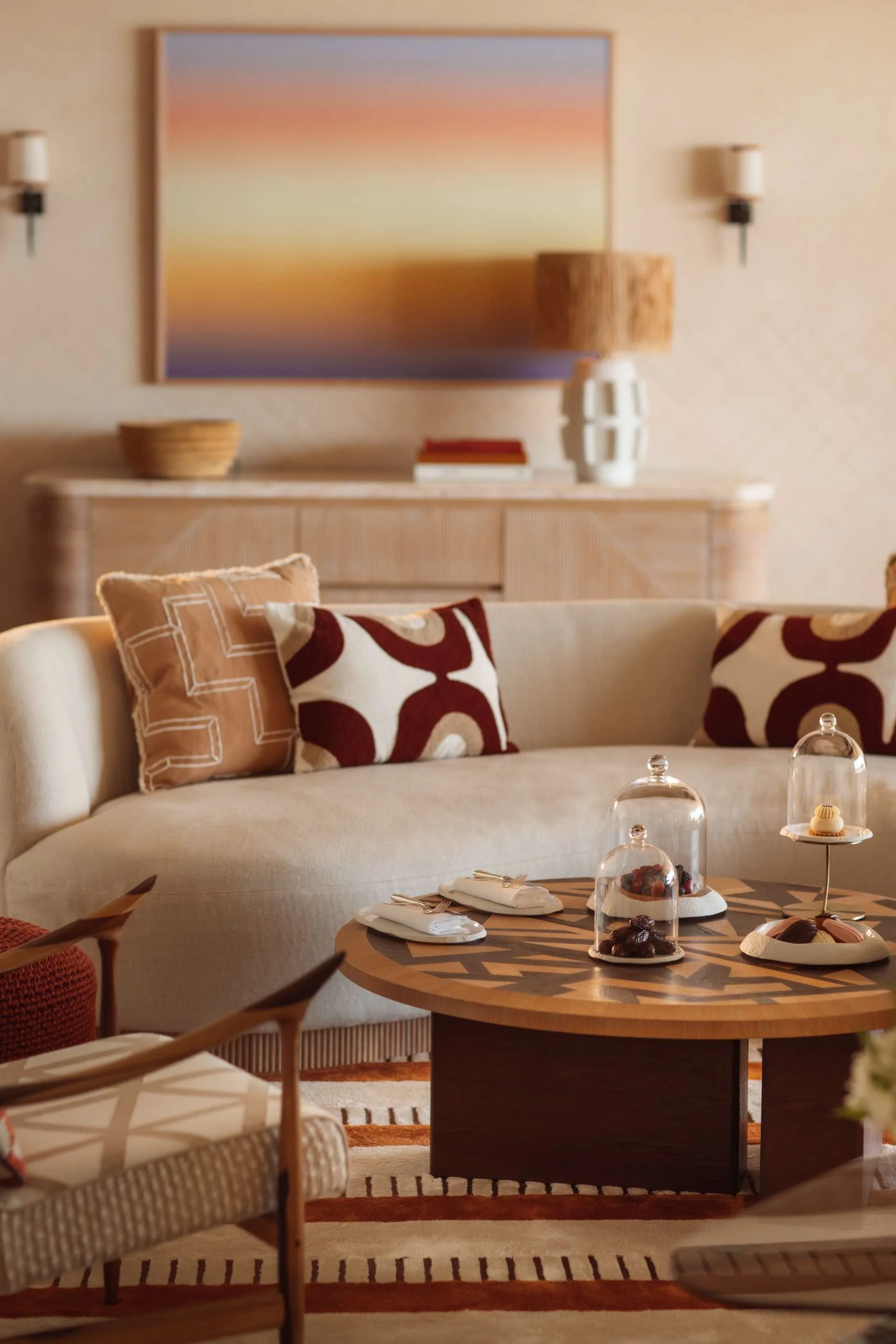 Living room with beige sofa, decorative pillows, wooden coffee table with glass cloches and desserts, patterned area rug, side chairs, a sideboard with a lamp and artwork, and wall sconces.