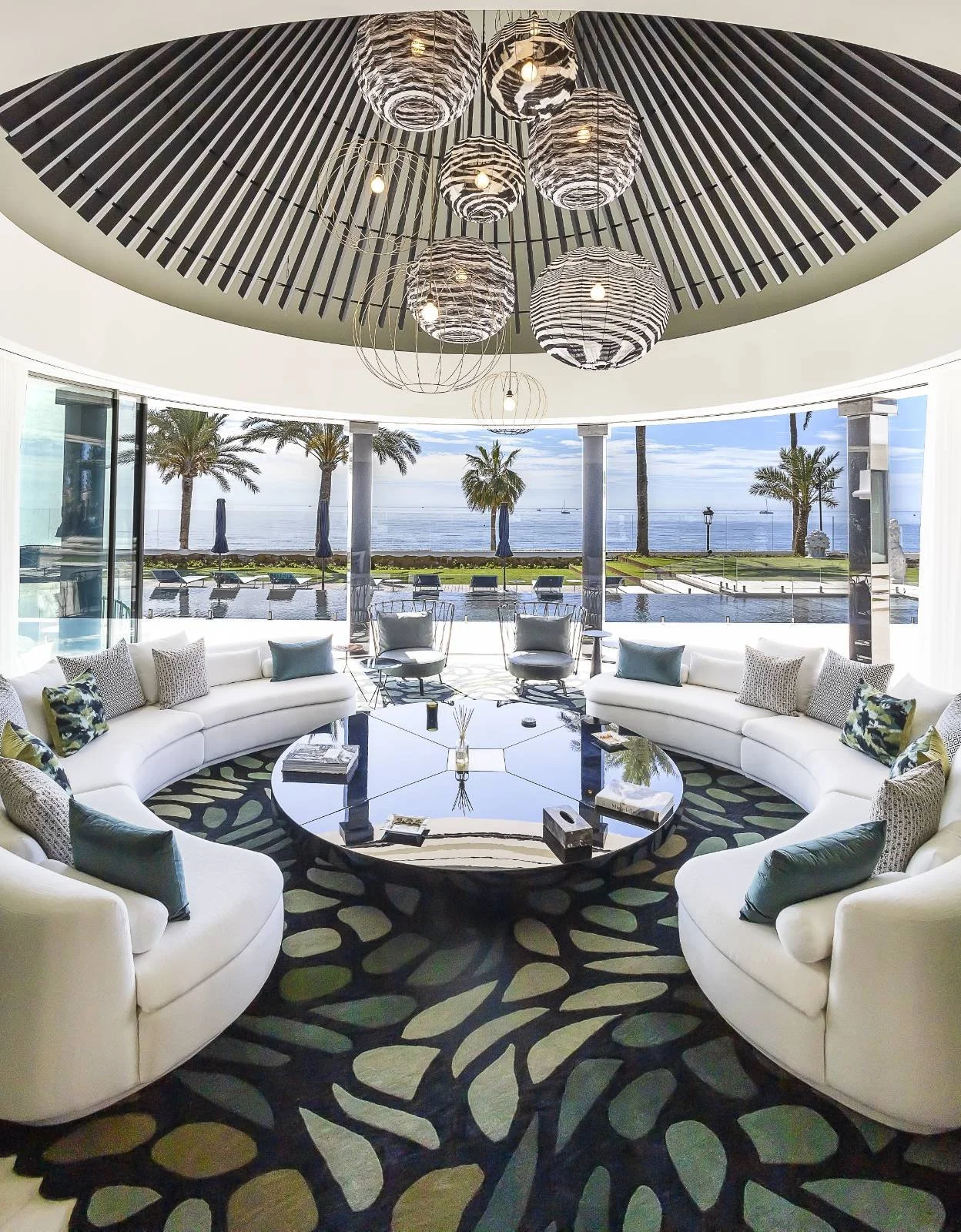 Modern lounge area with white curved sofa and an oval black coffee table, decorated with pillows, in front of large windows with a view of palm trees and a beach, with a circular ceiling featuring hanging spherical pendant lights.