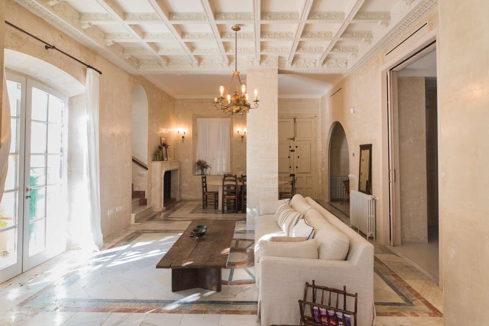 Bright living room with cream-colored walls, a white sofa, wooden coffee table, chandelier, and large windows with white curtains.