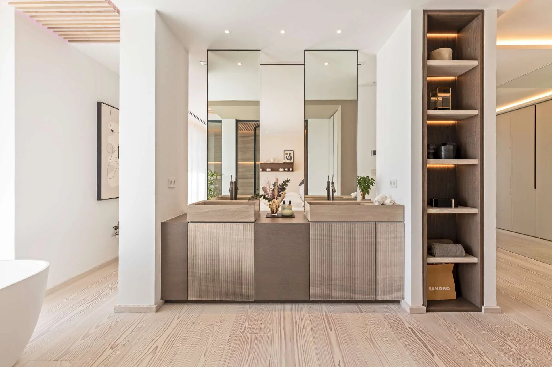 Modern bathroom with double sinks, large mirrors, light wood cabinetry, and a tall open shelving unit with decor and storage.