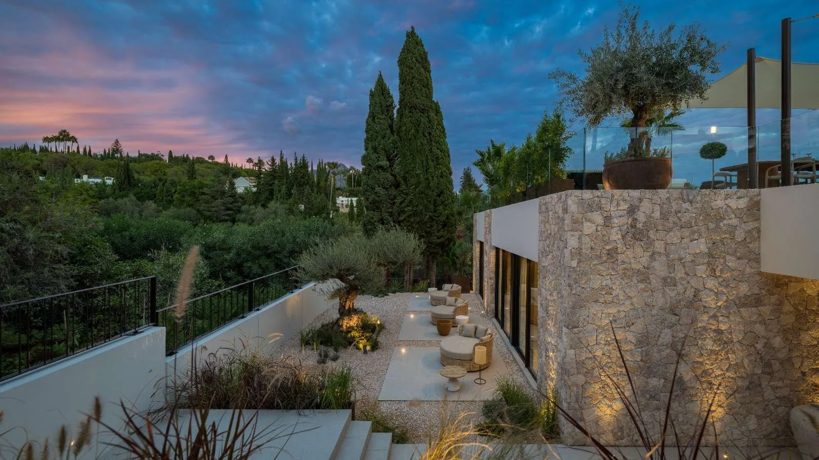 Luxury outdoor patio with modern furniture and lush greenery at dusk, overlooking a cityscape and colorful sky.