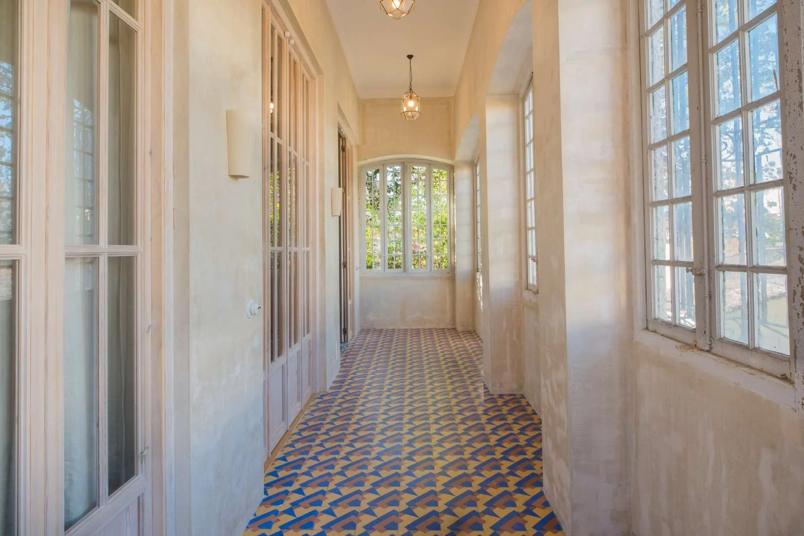 Bright enclosed porch with multiple windows, light-colored walls, hanging light fixtures, and a patterned tile floor.