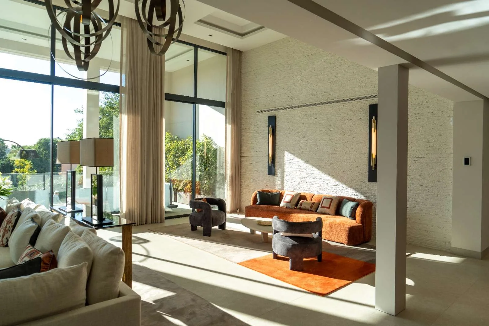 Modern living room with large floor-to-ceiling windows, beige curtains, a sofa, two armchairs, a coffee table, and decorative wall lamps, filled with natural sunlight.