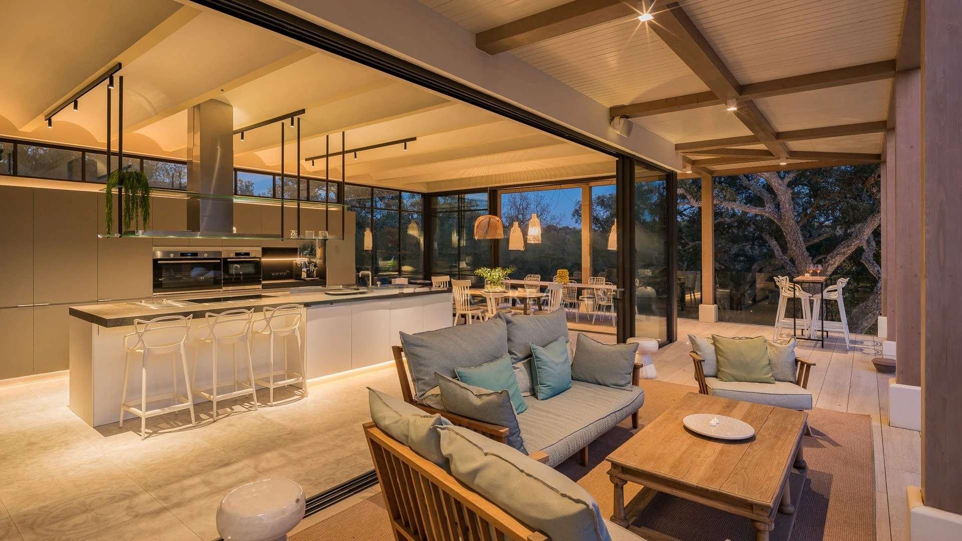 Open-concept living room and kitchen with wooden accents, large glass windows, and outdoor view of trees. Modern furniture, neutral tones, and cozy lighting create a warm atmosphere.