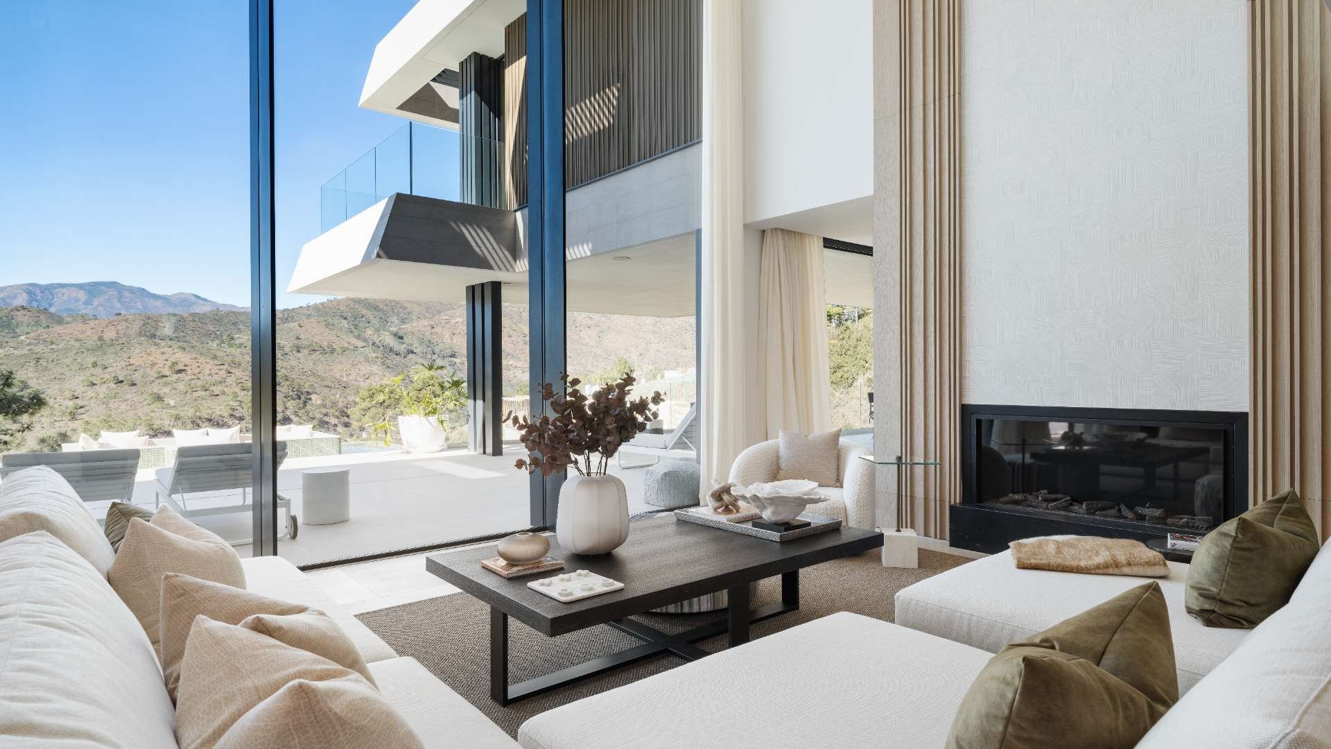 Modern living room with large glass sliding doors, mountain view, white sofa with beige and green pillows, central wooden coffee table with decorative items, fireplace, and a balcony with outdoor seating.