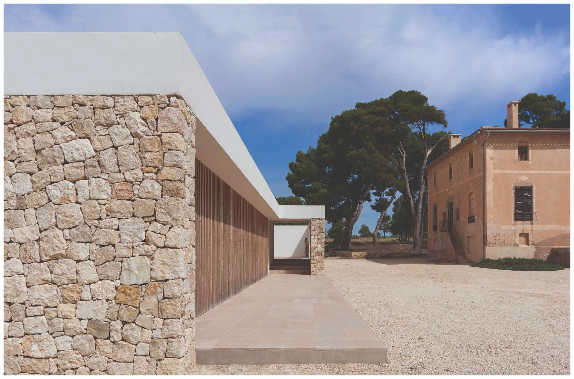 Modern building with stone and wood exterior, large white roof, and an open walkway, with an older peach-colored house and trees in the background.