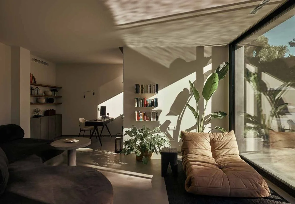Bright living room with large sliding glass door, tan lounge chair, green indoor plants, bookshelf, desk with computer, and modern decor.